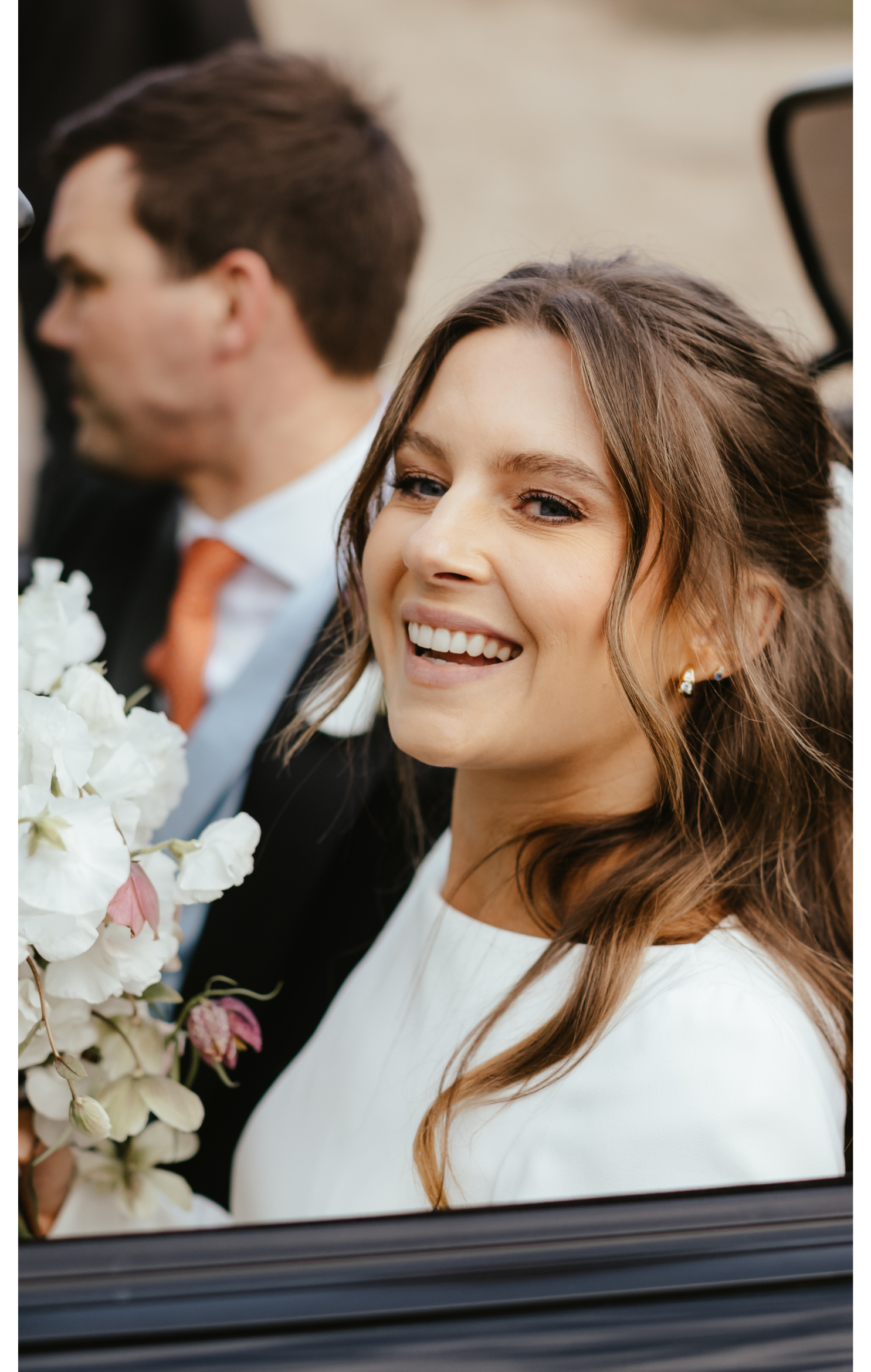 A smiling woman with long brown hair and pearl earrings, sitting in a car with a man in a tuxedo beside her, holding a bouquet of white flowers.