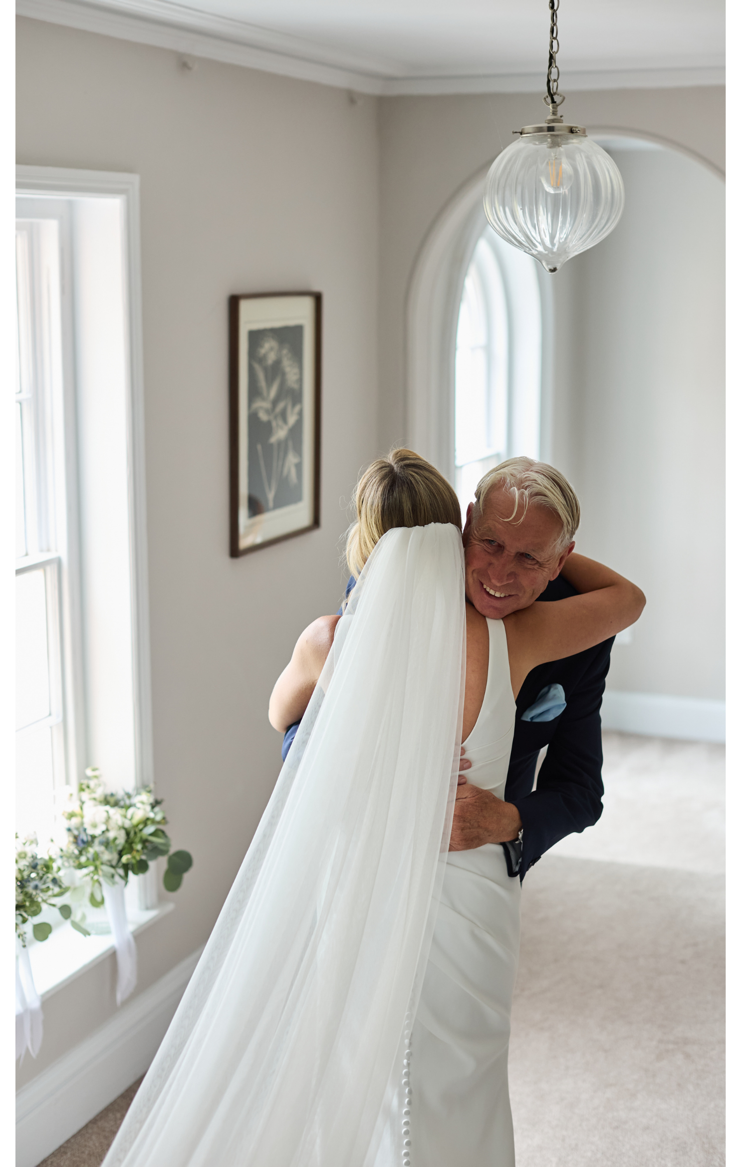 A bride hugging an older man, possibly her father, in a bright room with large windows and a hanging light fixture. The bride is wearing a white wedding dress and veil, and the man is in a dark suit.