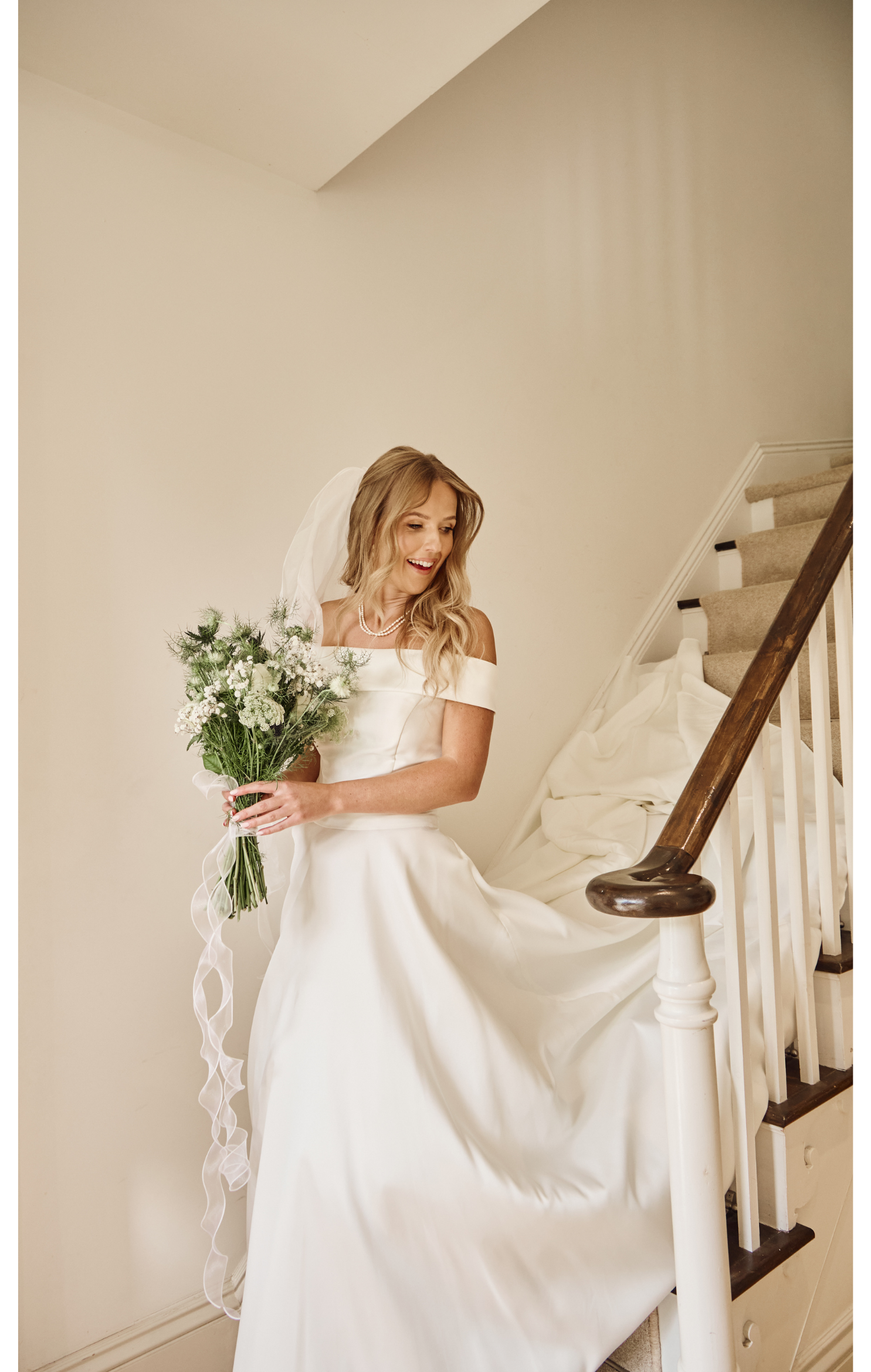 A bride in a white wedding gown holding a bouquet of flowers, standing on a staircase, smiling and looking down.