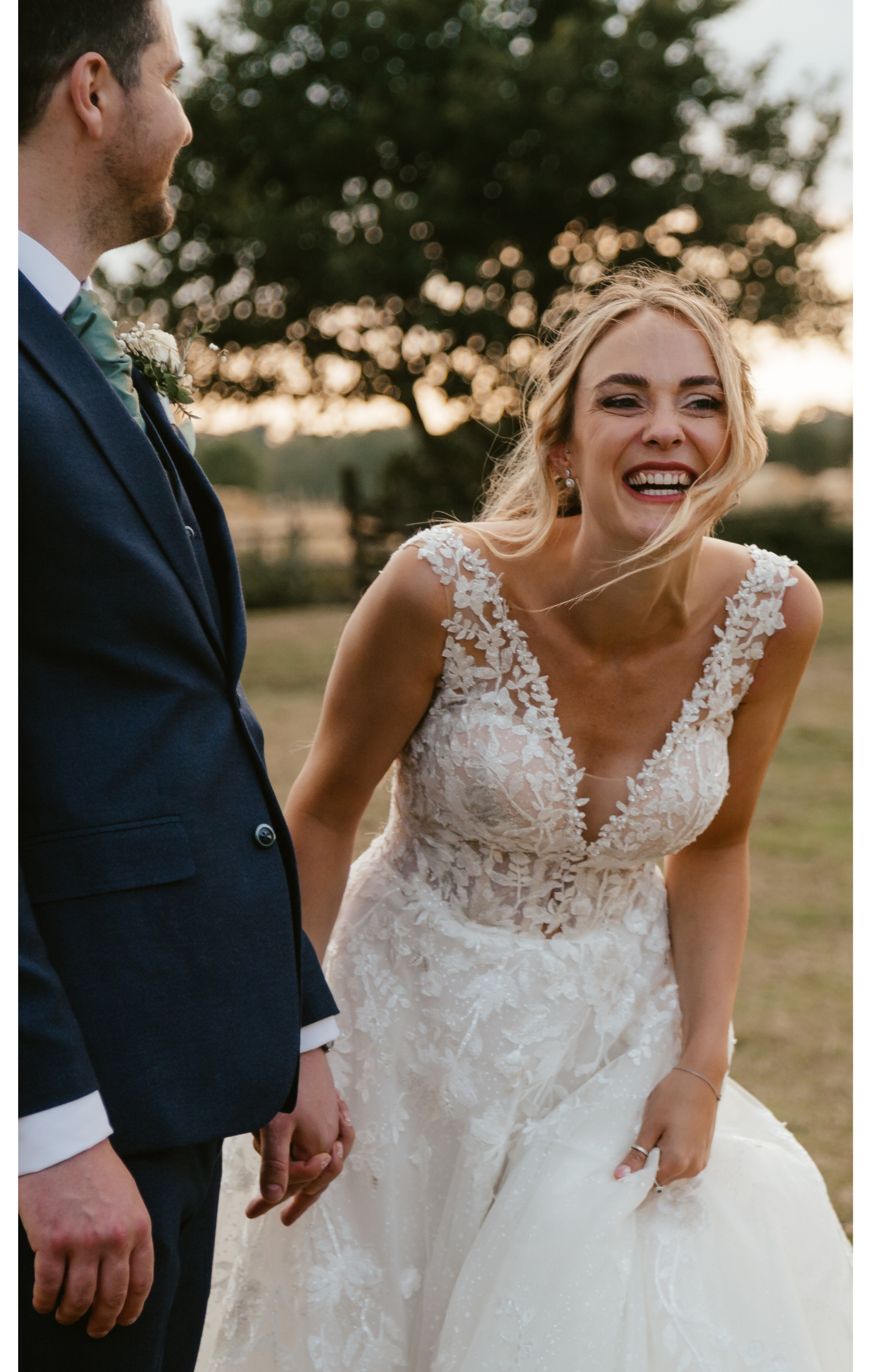 A bride laughing and holding her wedding dress while standing outdoors with a groom holding her hand.