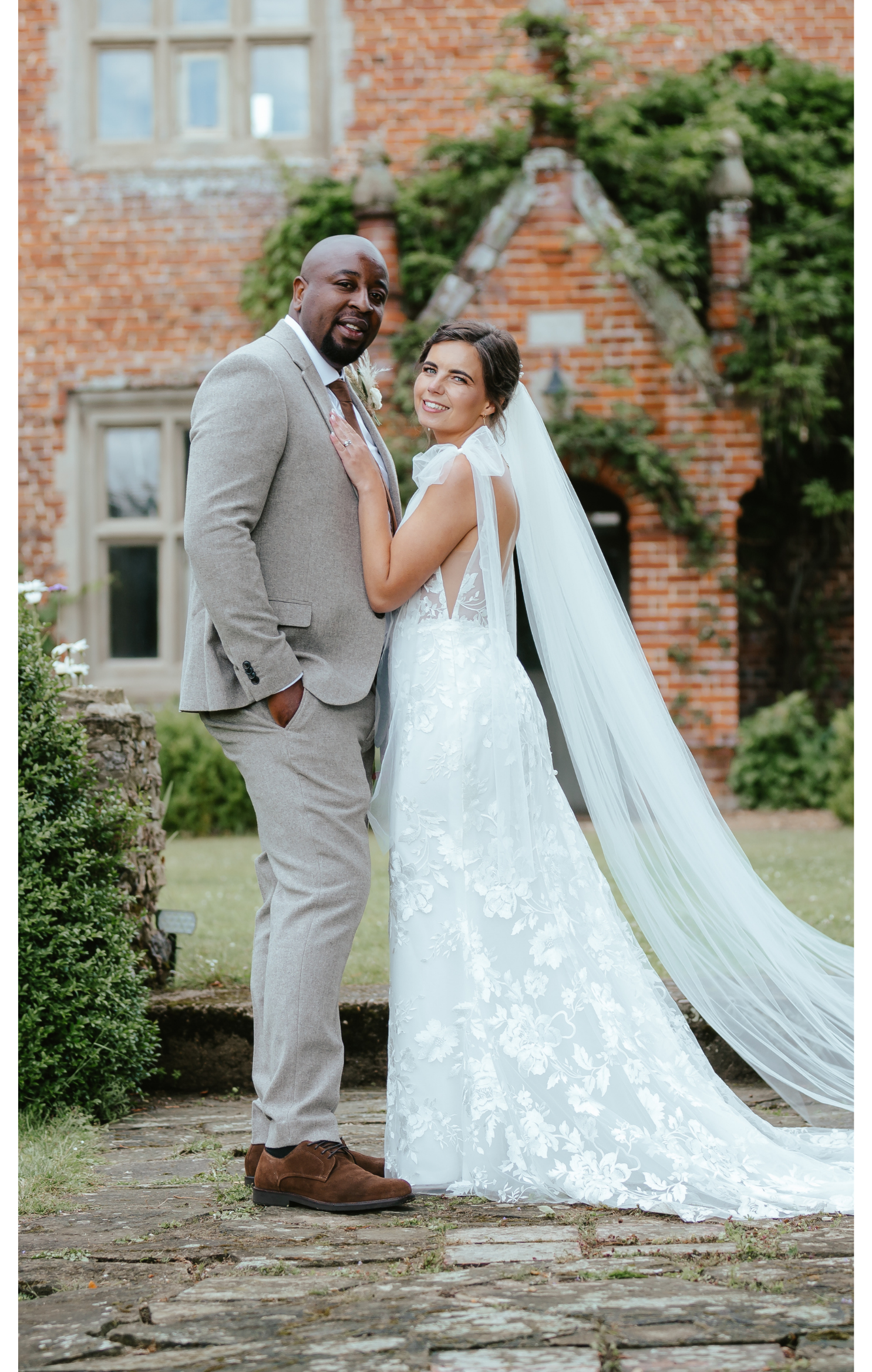 A bride and groom pose outdoors, with the bride in a white wedding gown and long veil, and the groom in a light gray suit. They stand on a stone path in front of a red-brick building with greenery.