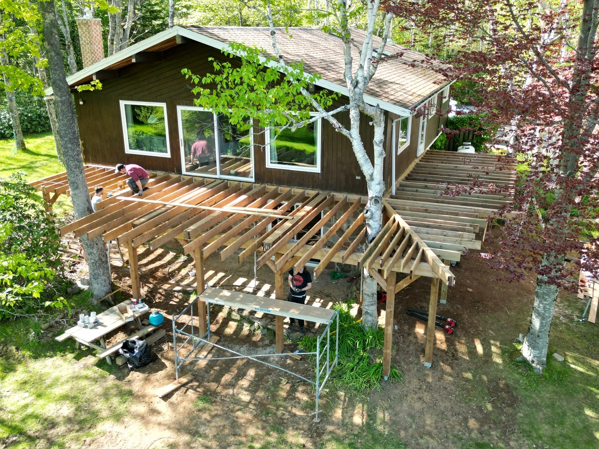 A house with a new wooden deck in construction surrounded by trees and greenery, with workers installing the deck, including framing and support beams.