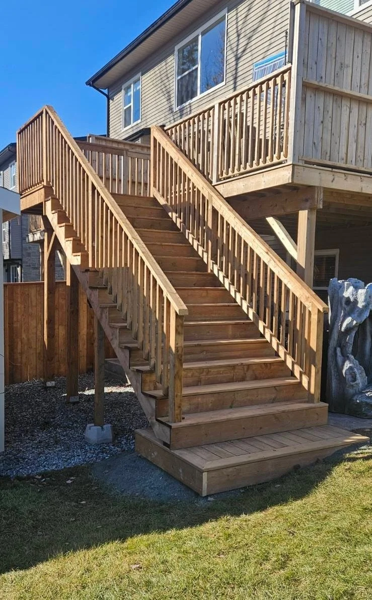 Newly built wooden backyard staircase leading up to a raised deck on a house, with a grassy yard in the foreground and a blue sky.