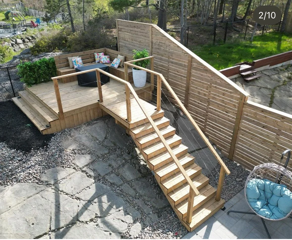 A newly built wooden deck with stairs and a privacy screen, furnished with outdoor seating and decorative pillows, surrounded by yard and garden.