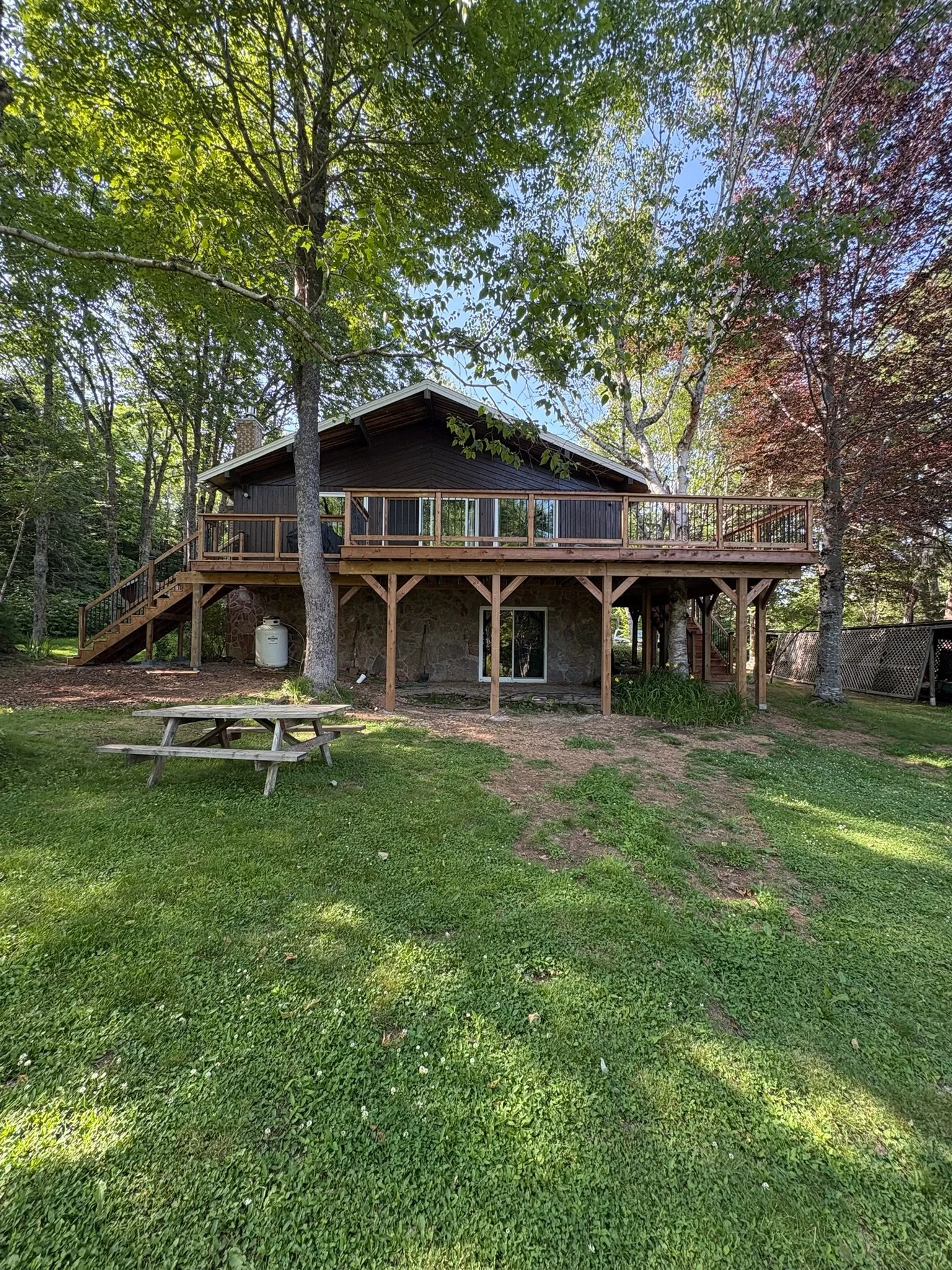 A house with a large wooden deck and stairs in a backyard surrounded by trees and green grass.