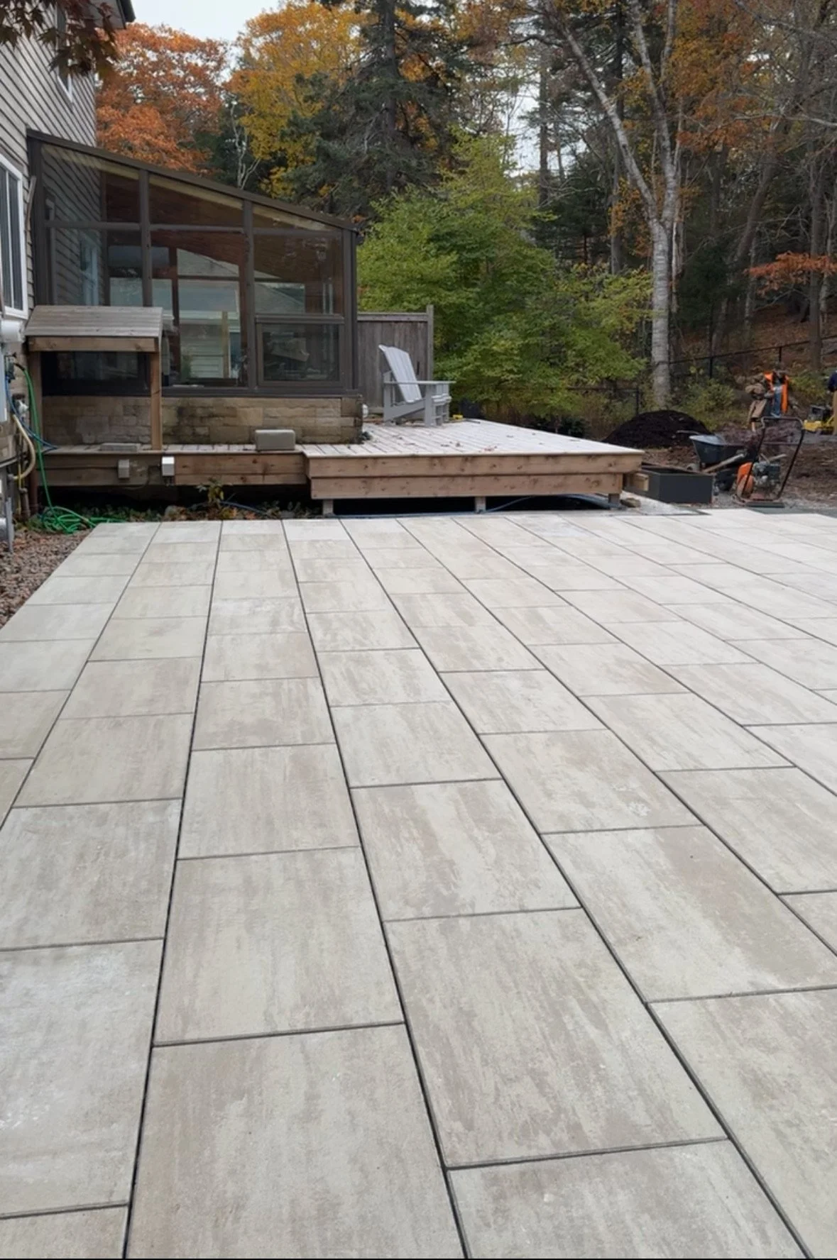 A backyard patio with newly installed light-colored rectangular tiles, a raised wooden deck with a screened porch, a white Adirondack chair, and surrounding trees with colorful autumn leaves.