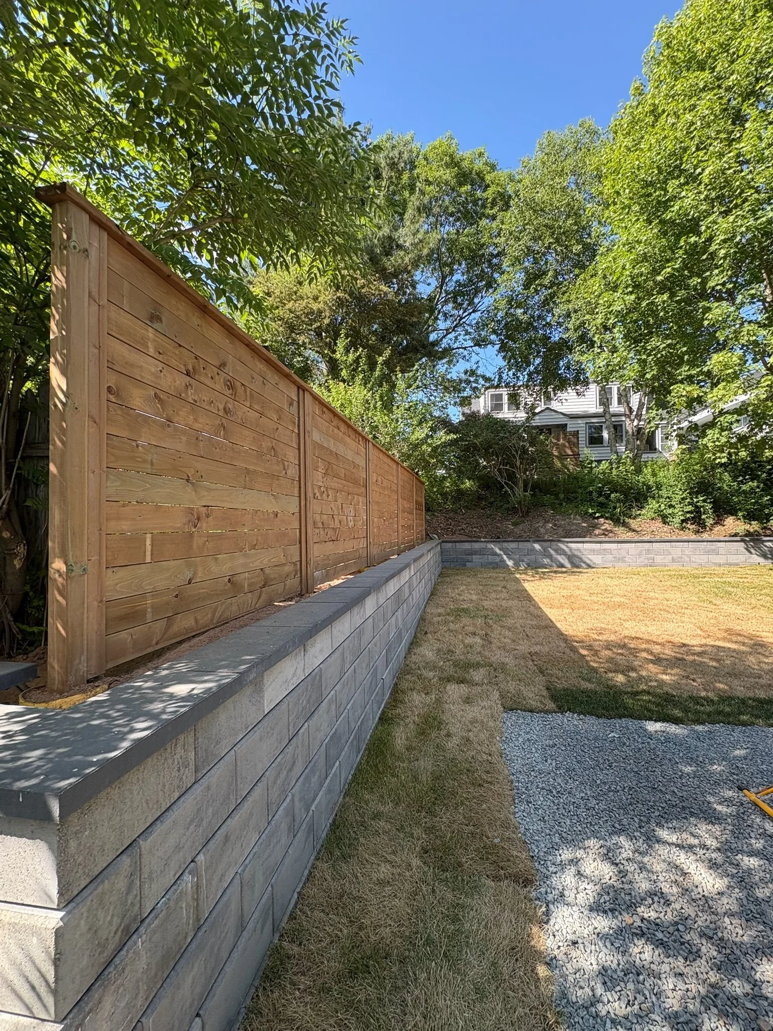A backyard with a newly constructed wooden fence, a low gray stone wall, a grass lawn, and trees under a clear blue sky.