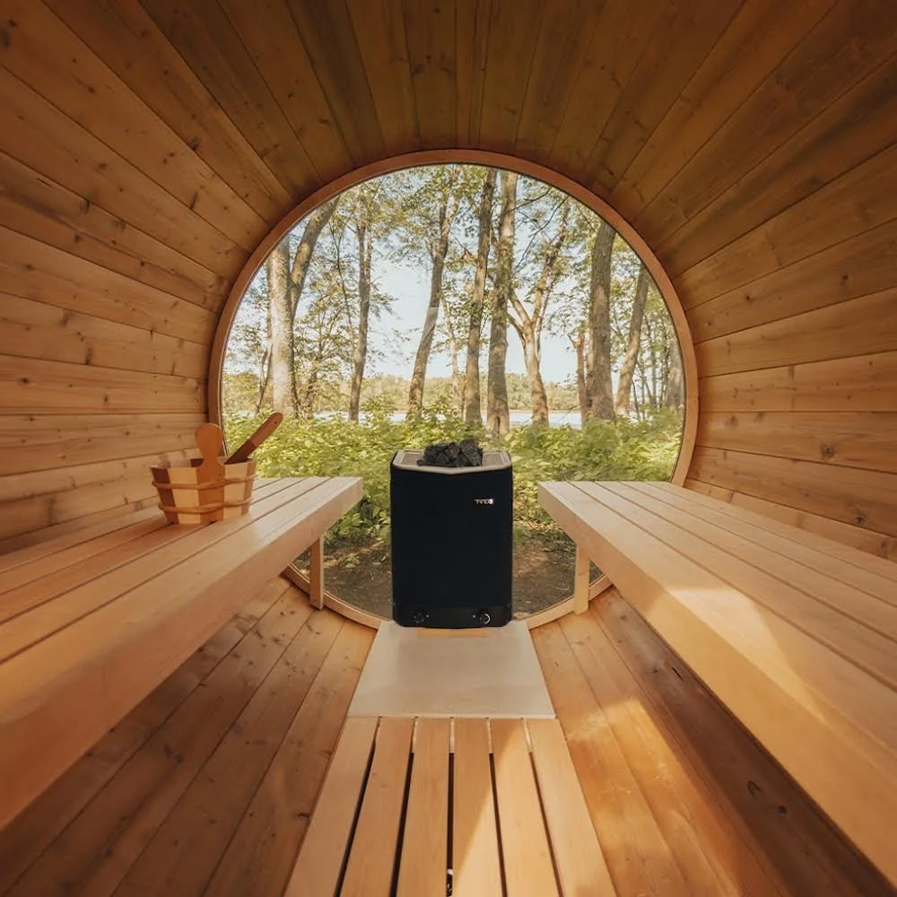 Interior of a wooden barrel sauna with a large circular window overlooking trees and a body of water. There are two wooden benches, a sauna bucket with a ladle on the left bench, and a black heater in the center with sauna stones on top.