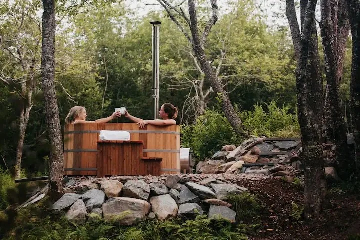 Two women relaxing in a wooden hot tub outdoors surrounded by trees and rocks, holding mugs and enjoying nature.