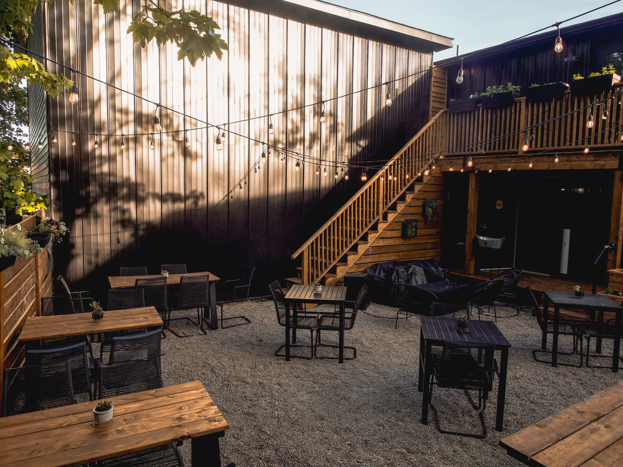 Outdoor patio with wooden tables and black chairs, string lights hanging overhead, and a staircase leading to an upper deck, surrounded by wooden fencing and greenery.