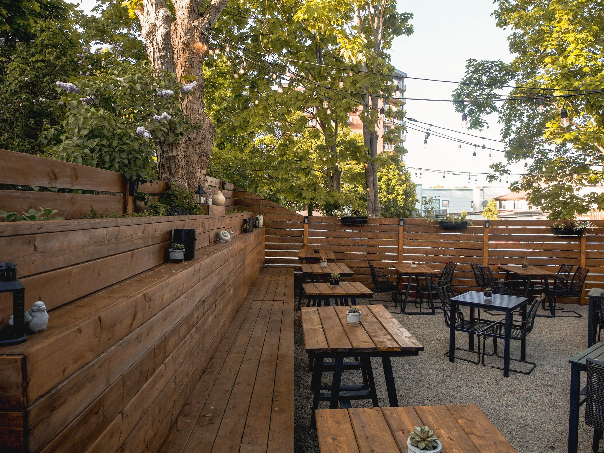 Outdoor patio with wooden tables and black chairs, surrounded by a wooden fence, with string lights hanging overhead and trees providing shade.