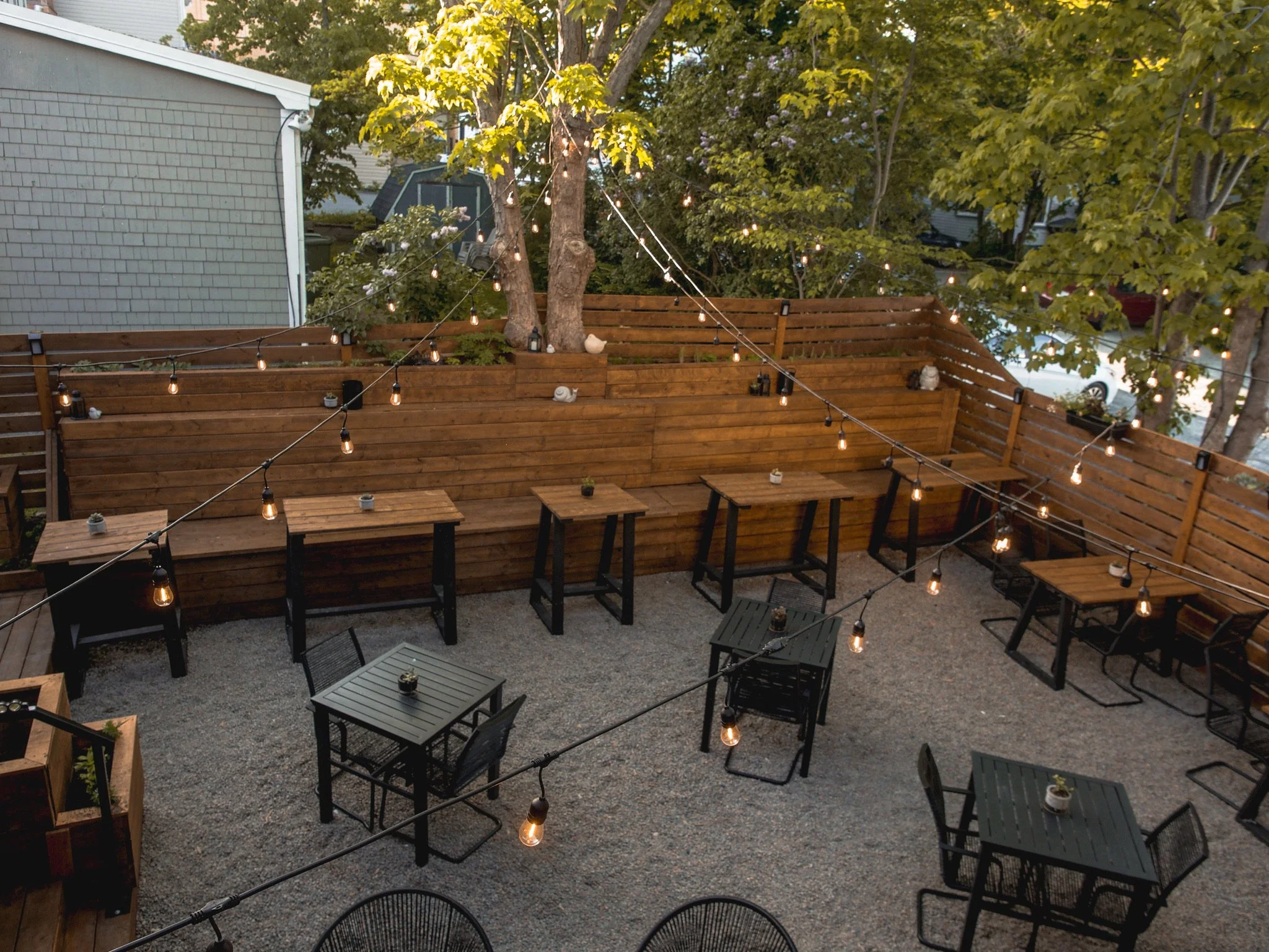 An outdoor patio area with several black tables and chairs, string lights overhead, and a wooden privacy fence, surrounded by trees.