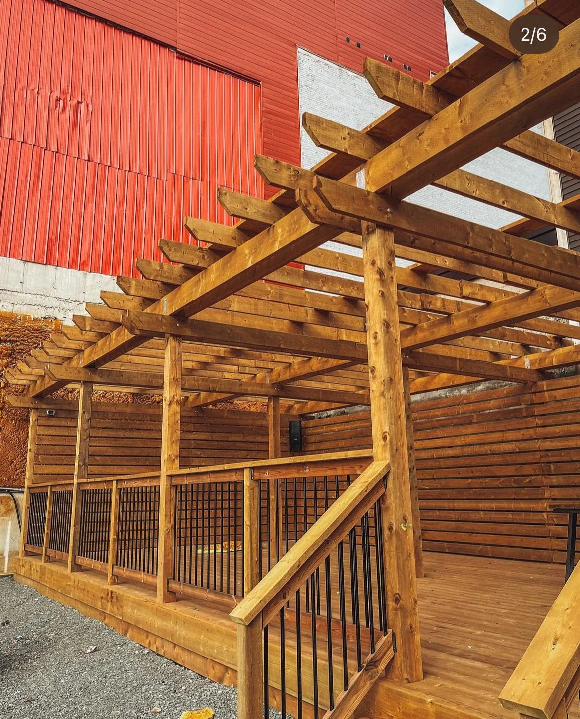 A wooden deck with a pergola and black metal railing in front of a modern building with red siding and white brick accents.