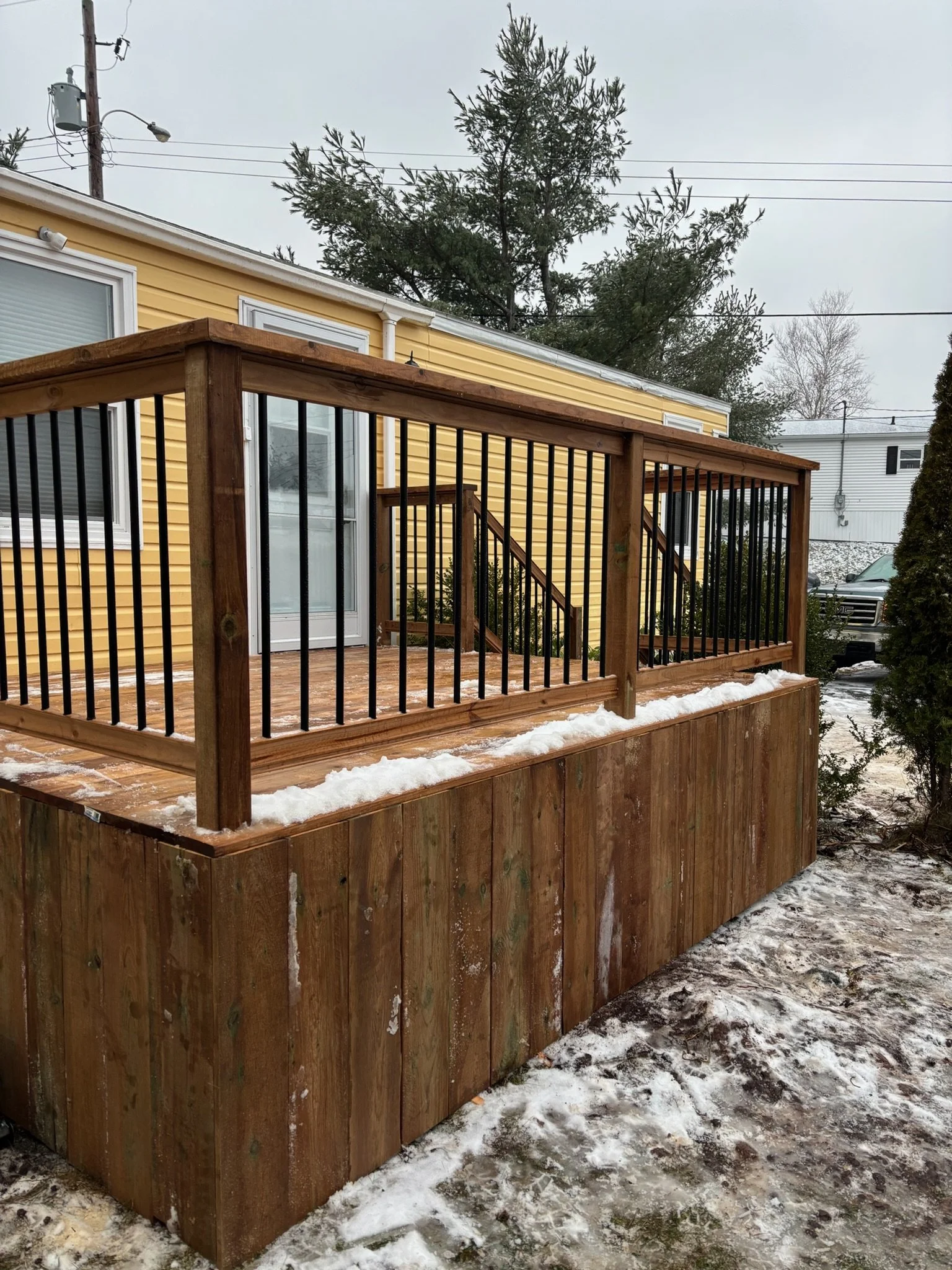 Wooden deck attached to yellow house with snow on top and surrounding ground, with black metal railing, in a residential area with trees and parked cars.
