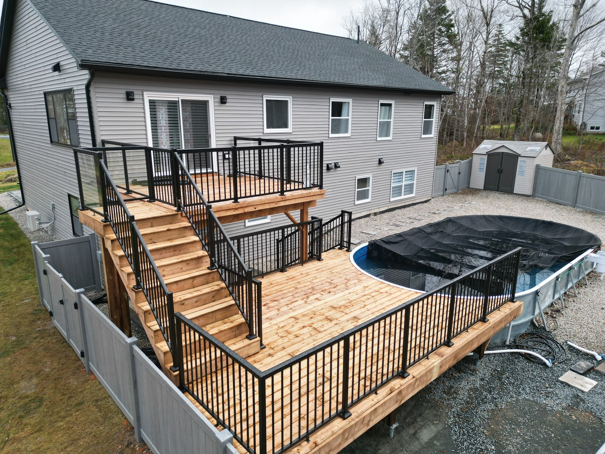 A newly constructed wooden deck attached to a gray house, featuring black metal railings, stairs leading down to the yard, and a swimming pool covered with a black tarp, surrounded by gravel and a gray fence.