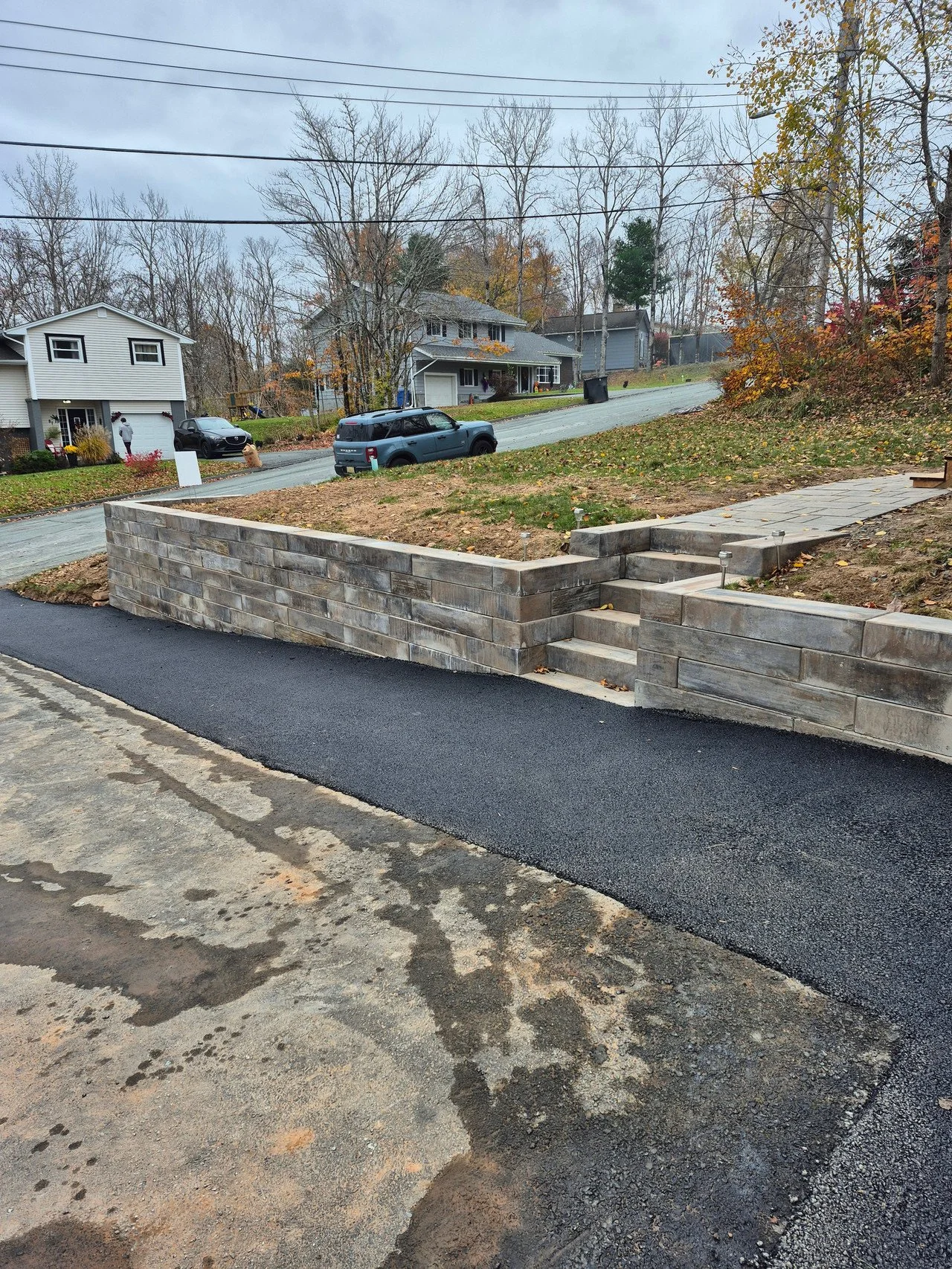 A residential street with houses, parked cars, a landscaped yard with a new stone retaining wall and stairs, and trees with autumn foliage.