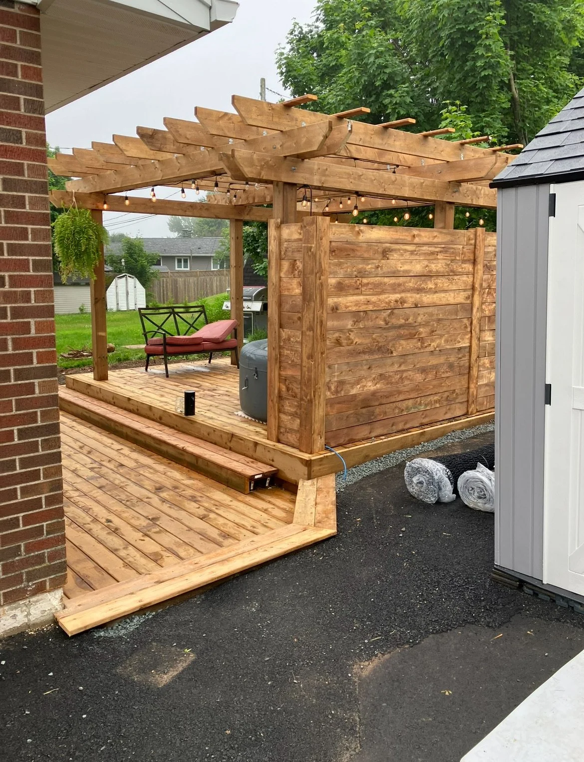 A newly constructed wooden backyard pergola with string lights, a deck, and outdoor seating, situated beside a brick house and a shed, with a green lawn and trees in the background.