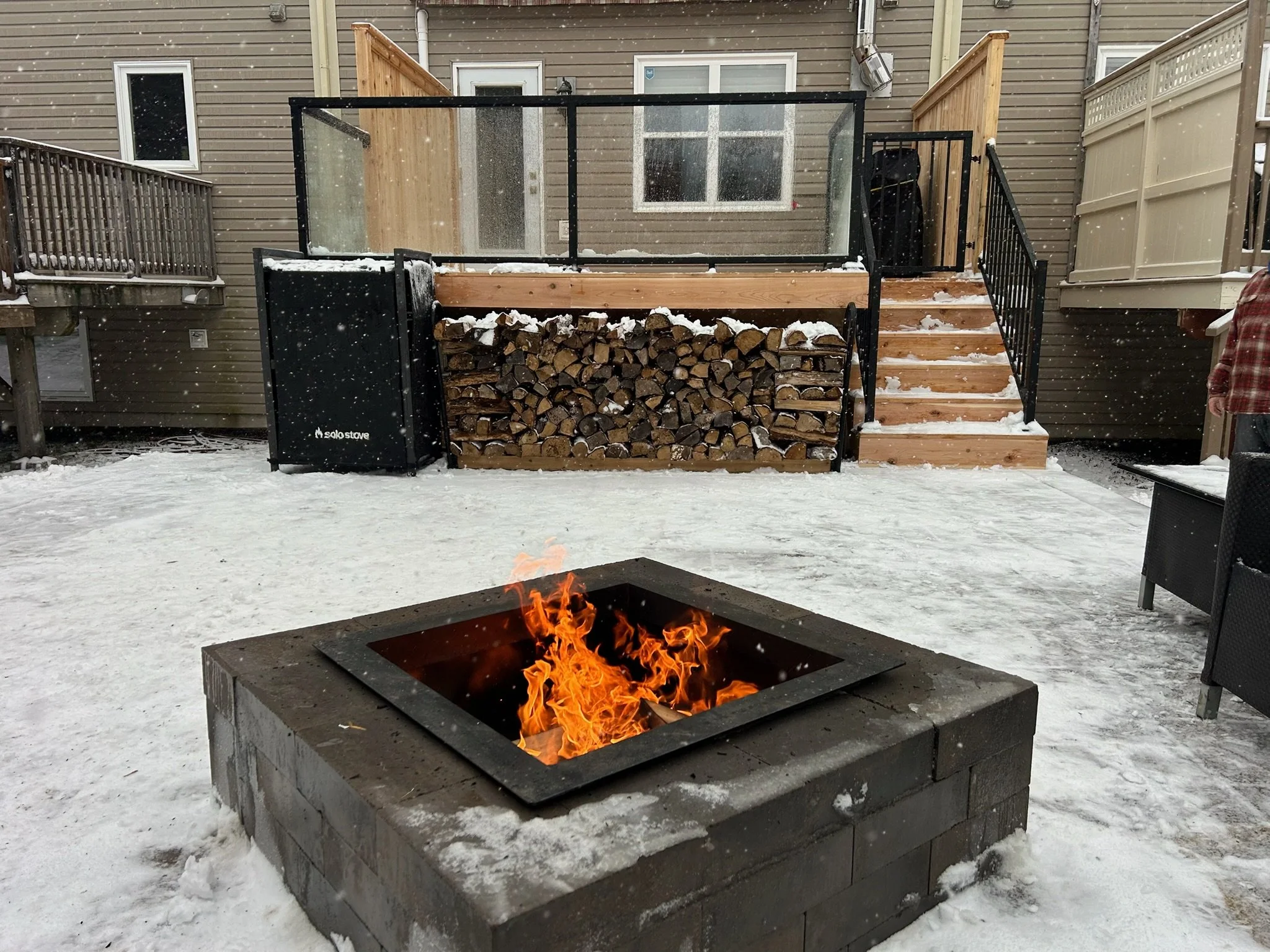 Snow-covered backyard with a fire pit in the foreground and a raised deck with woodpile in the background.