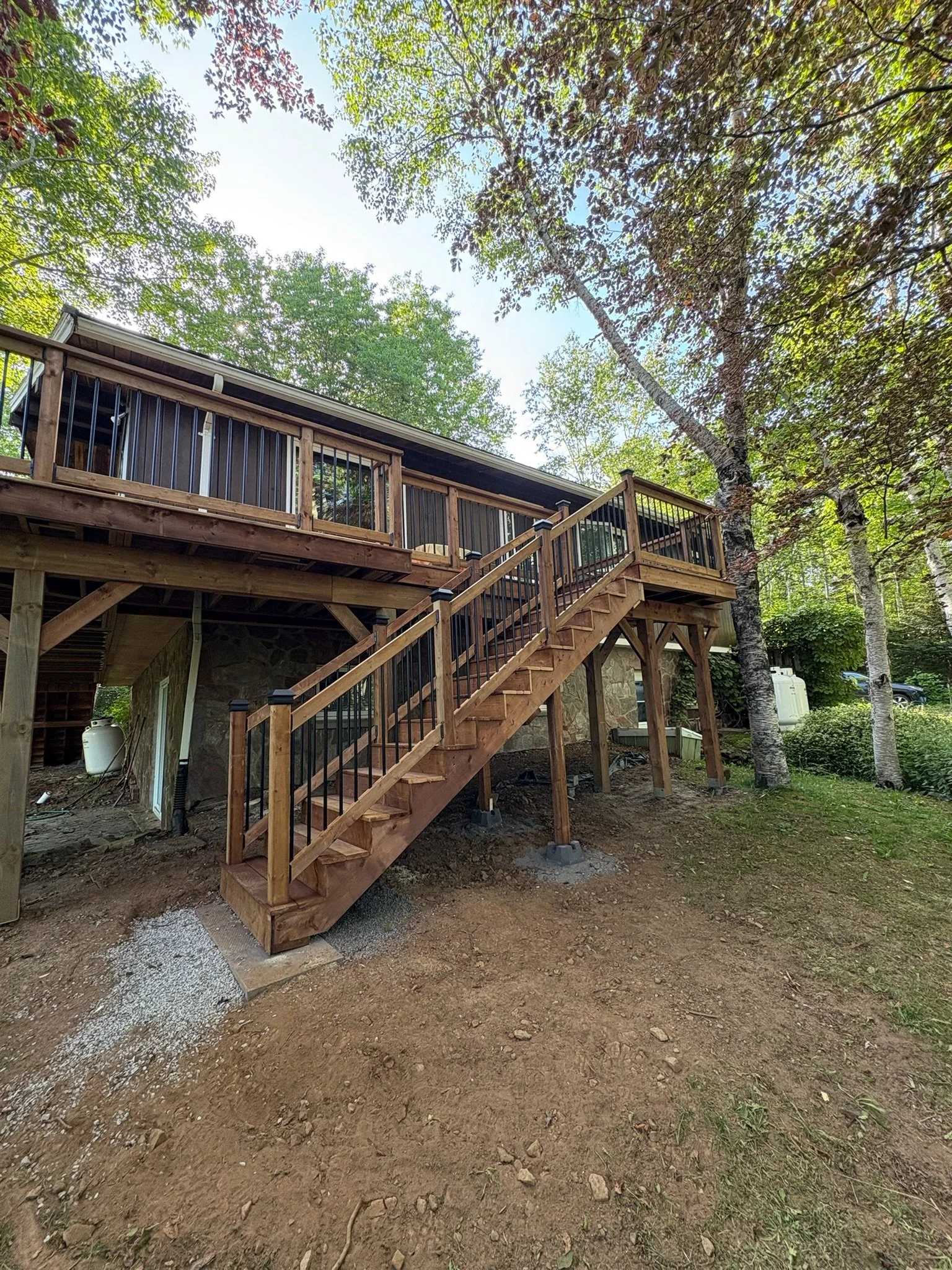 A newly built wooden staircase with black railing leading up to a house deck surrounded by trees and greenery during daytime.
