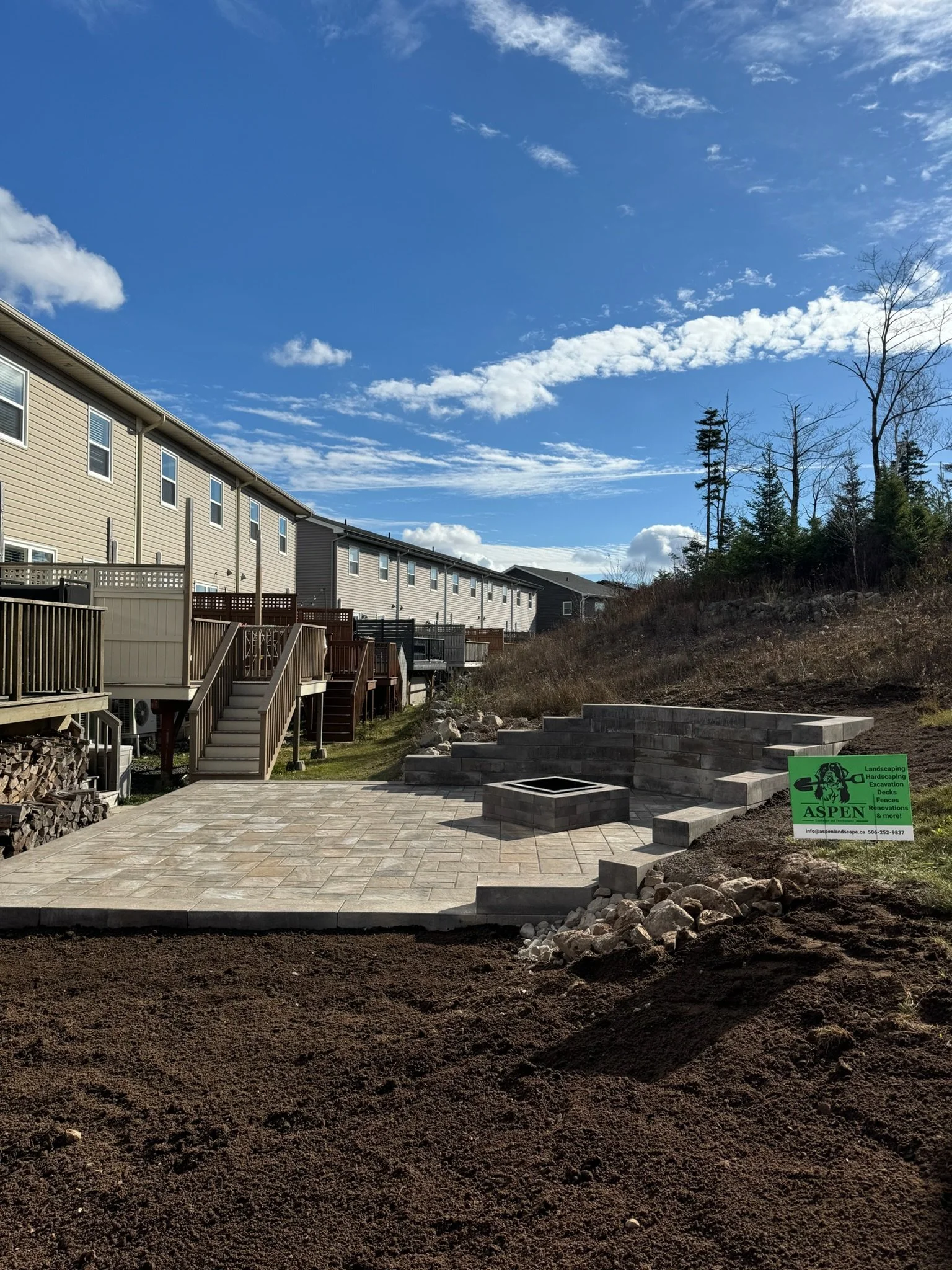 A newly constructed outdoor patio with stone pavers, staircase, and fire pit area, adjacent to a hillside with grass and trees. Multi-story houses with decks are visible in the background, under a partly cloudy blue sky.