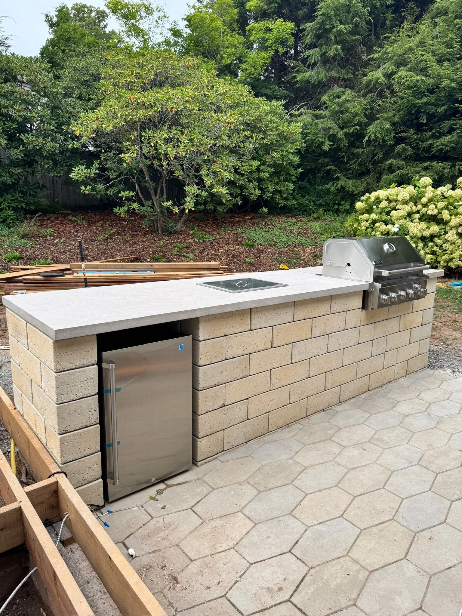 Outdoor kitchen with a brick counter, a built-in grill, a small refrigerator, and a cooktop, set on a patio with hexagonal tiles, surrounded by green trees and bushes.