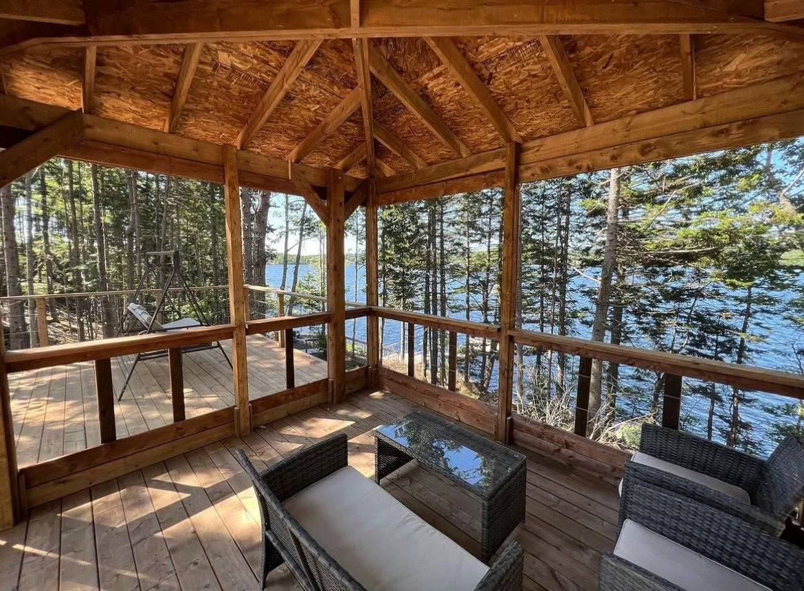 A wooden screened porch overlooking a forested lakeside view with trees and water visible in the background. There is outdoor furniture including a glass-top coffee table, rattan chairs with cushions, and a metal swing on the deck.