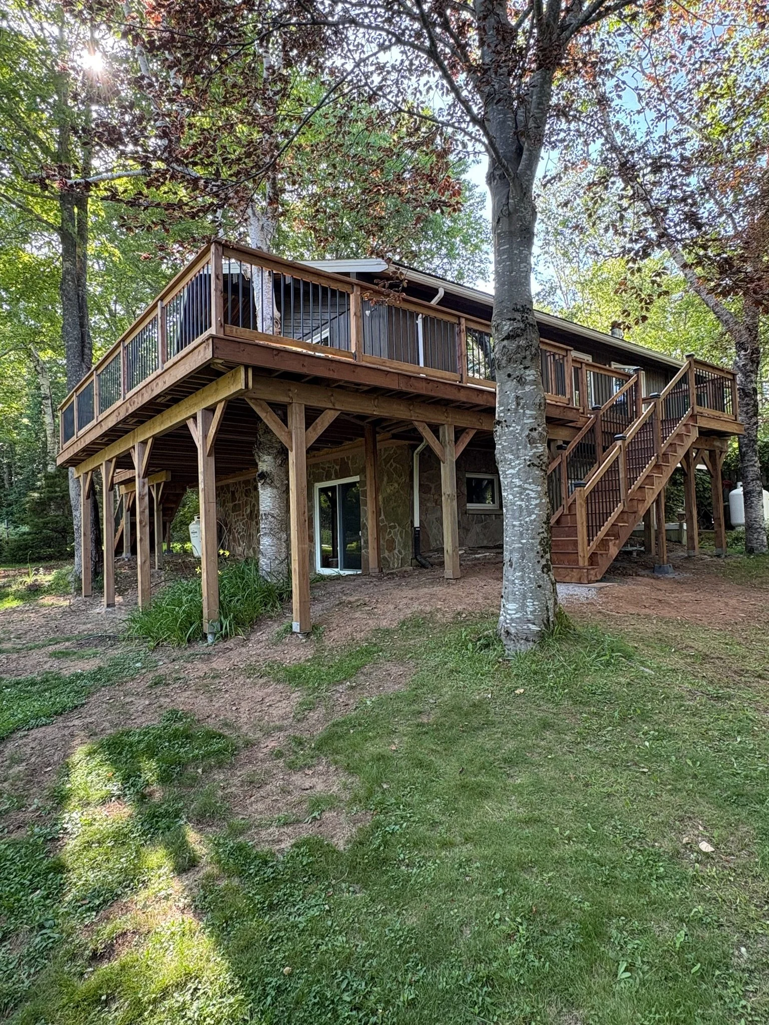 A two-story house in a wooded area with a large wooden deck and staircase on the right side, supported by wooden and stone pillars, with trees and greenery surrounding it.