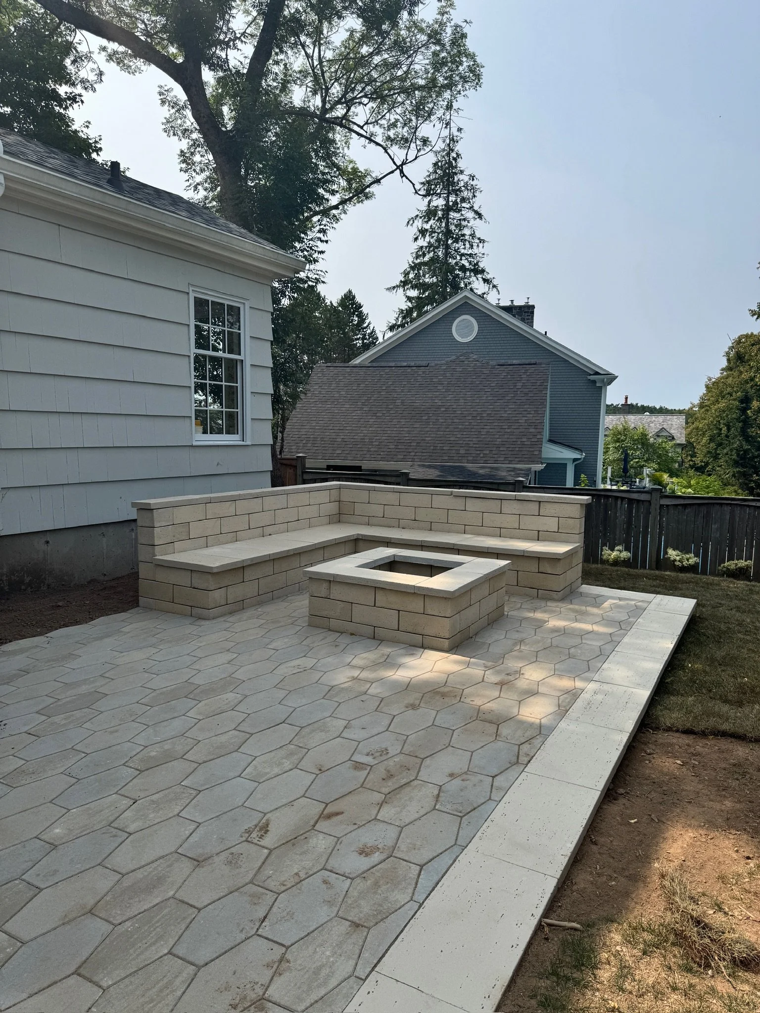 Newly constructed backyard patio with stone paving, built-in bench seating, and a fire pit, adjacent to a white house with a window and trees in the background.