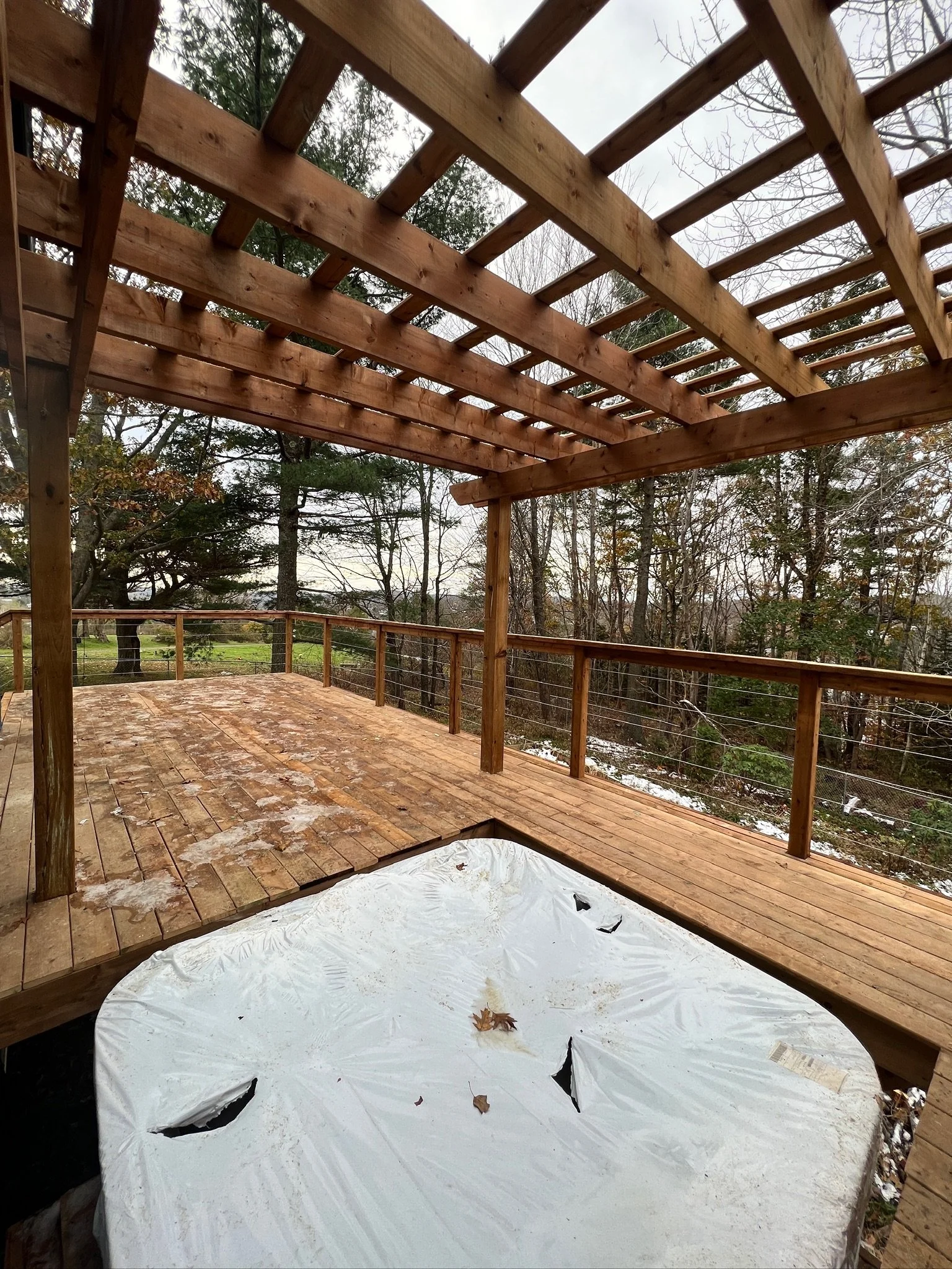 Wooden deck under construction with a partially built pergola. The deck has some water and leaves, and there is a covered hot tub in the bottom corner. Trees and a cloudy sky are in the background.