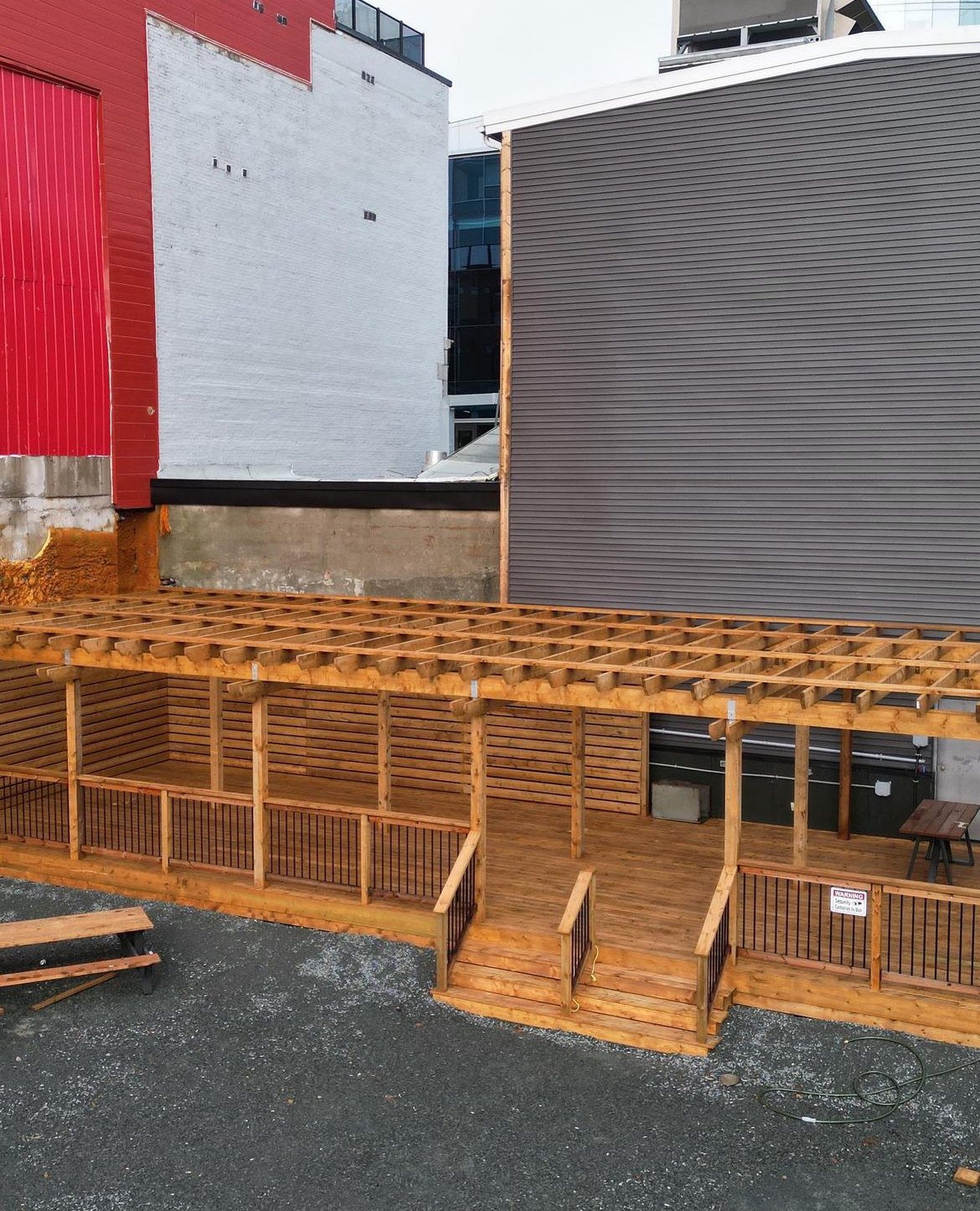 Construction site with a wooden deck and pergola, surrounded by buildings with various exterior finishes.
