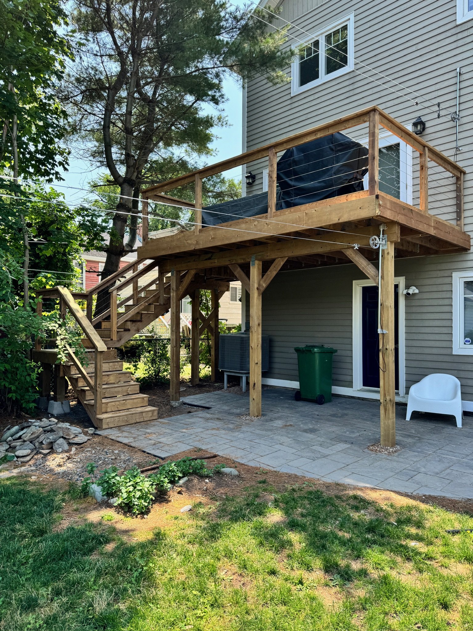 Backyard with a newly built wooden deck with stairs, stairs, a green trash bin, and paving stones, attached to a gray house with white trim and multiple windows.