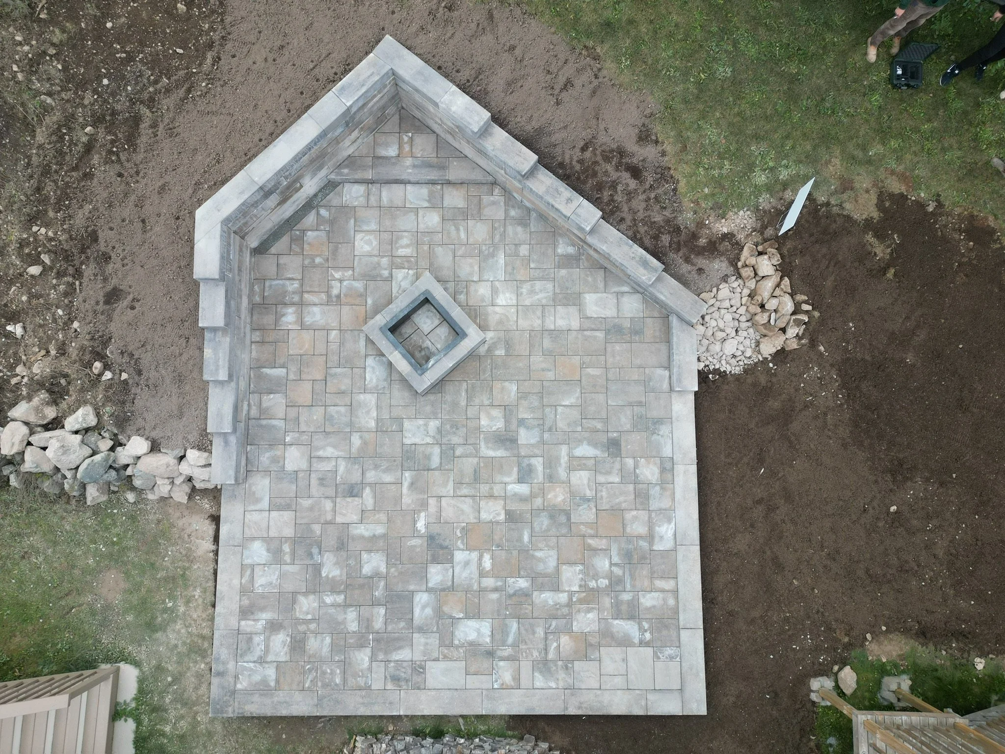 A partially constructed patio with stone pavers and a central square feature, surrounded by dirt and some grass, with construction tools and materials nearby.