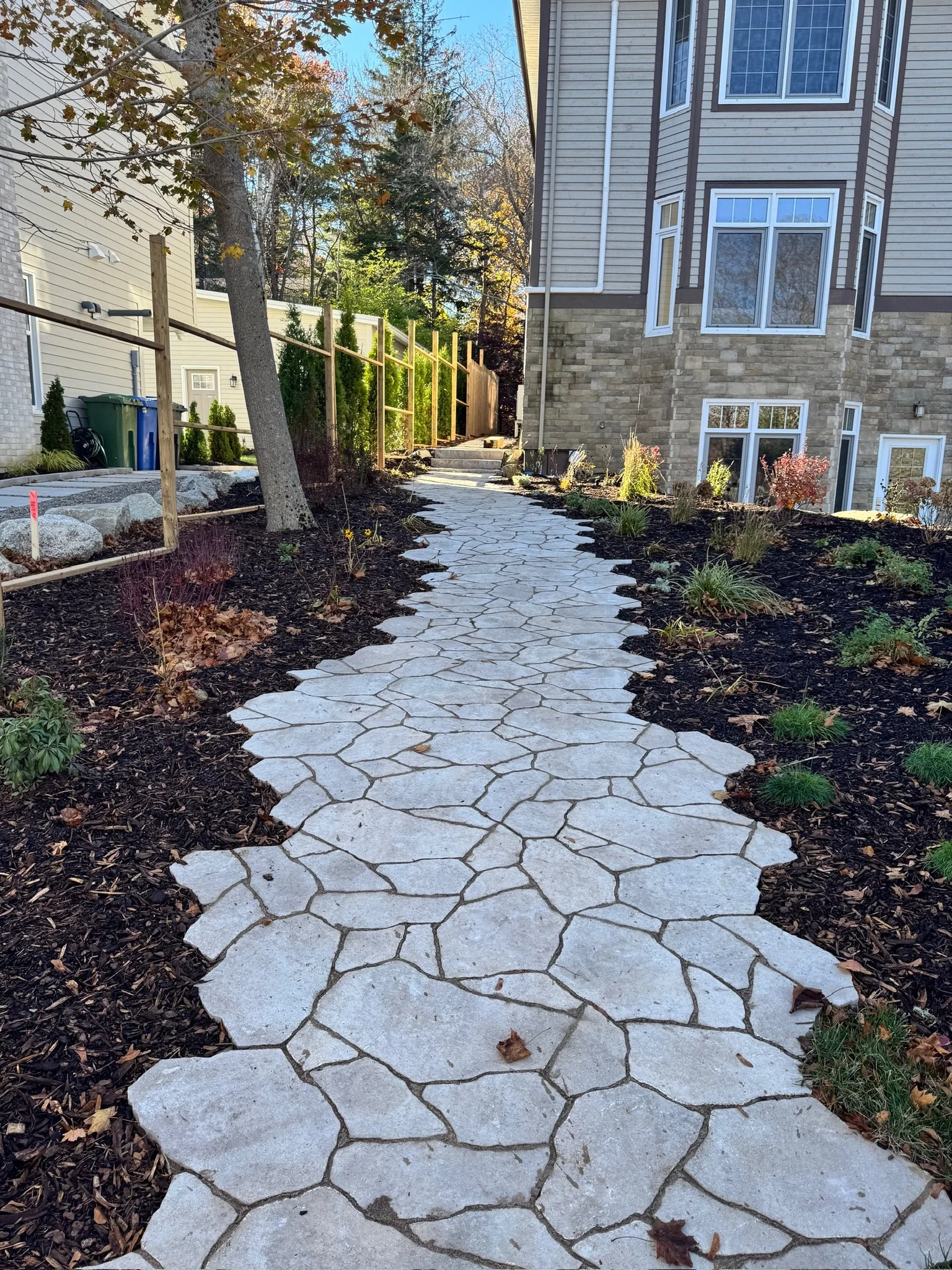 Stone walkway leading to stairs in backyard, flanked by garden beds with mulch and plants, and a wooden fence along the left side, adjacent to a house with beige siding and large windows.