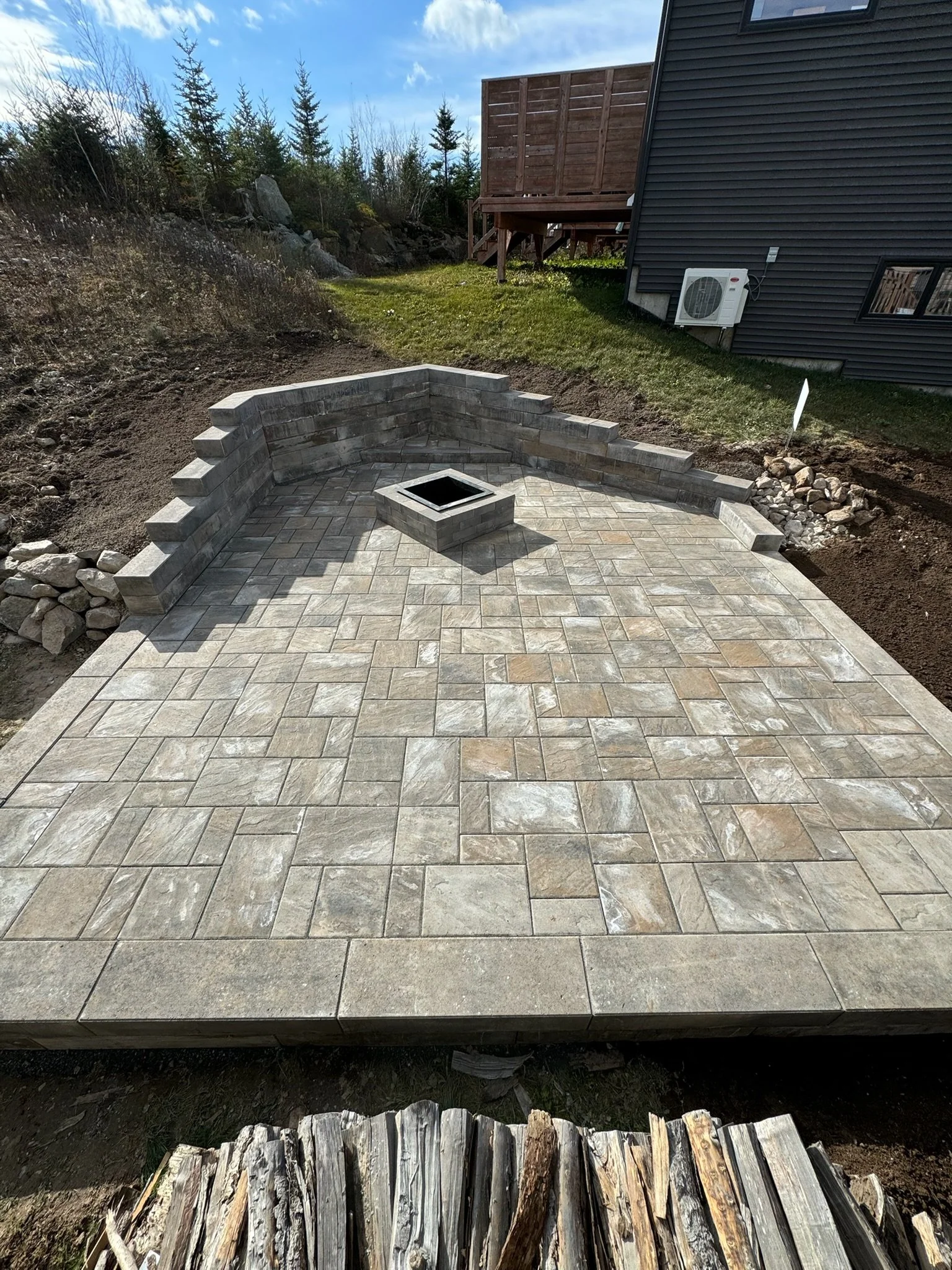Newly constructed outdoor patio with stone pavers, a fire pit, and a brick wall, situated behind a house with a black exterior, air conditioning unit, and a wooden deck, with trees and a cloudy sky in the background.