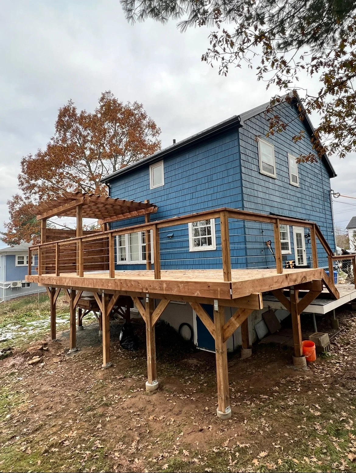 Newly built wooden deck with railing attached to a blue house, with a small covered pergola on the left side.