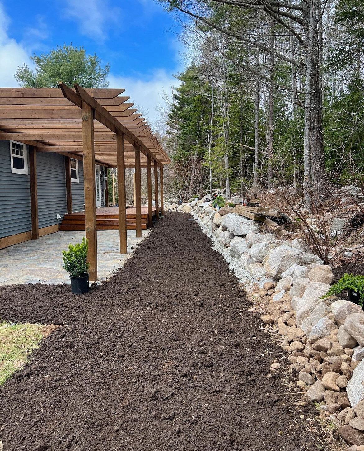 A backyard with a newly poured dirt pathway along the house, a small potted shrub at the corner, and a stone patio area. The house has blue siding and a wooden pergola extending from the back. There are large rocks and trees along the edge of the yar