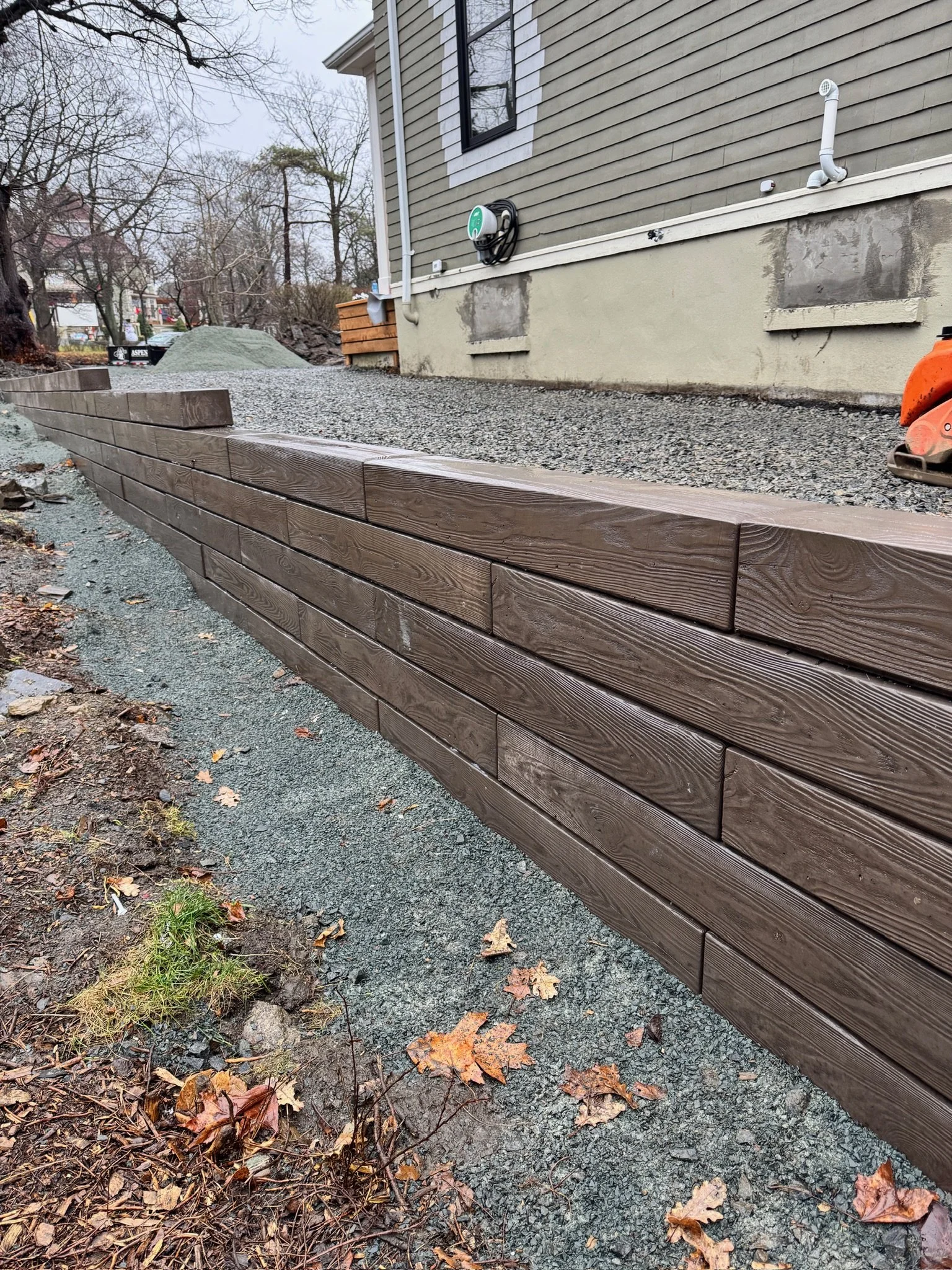 A residential construction site showing a new wooden retaining wall with a house in the background, construction materials, and fallen autumn leaves on the ground.
