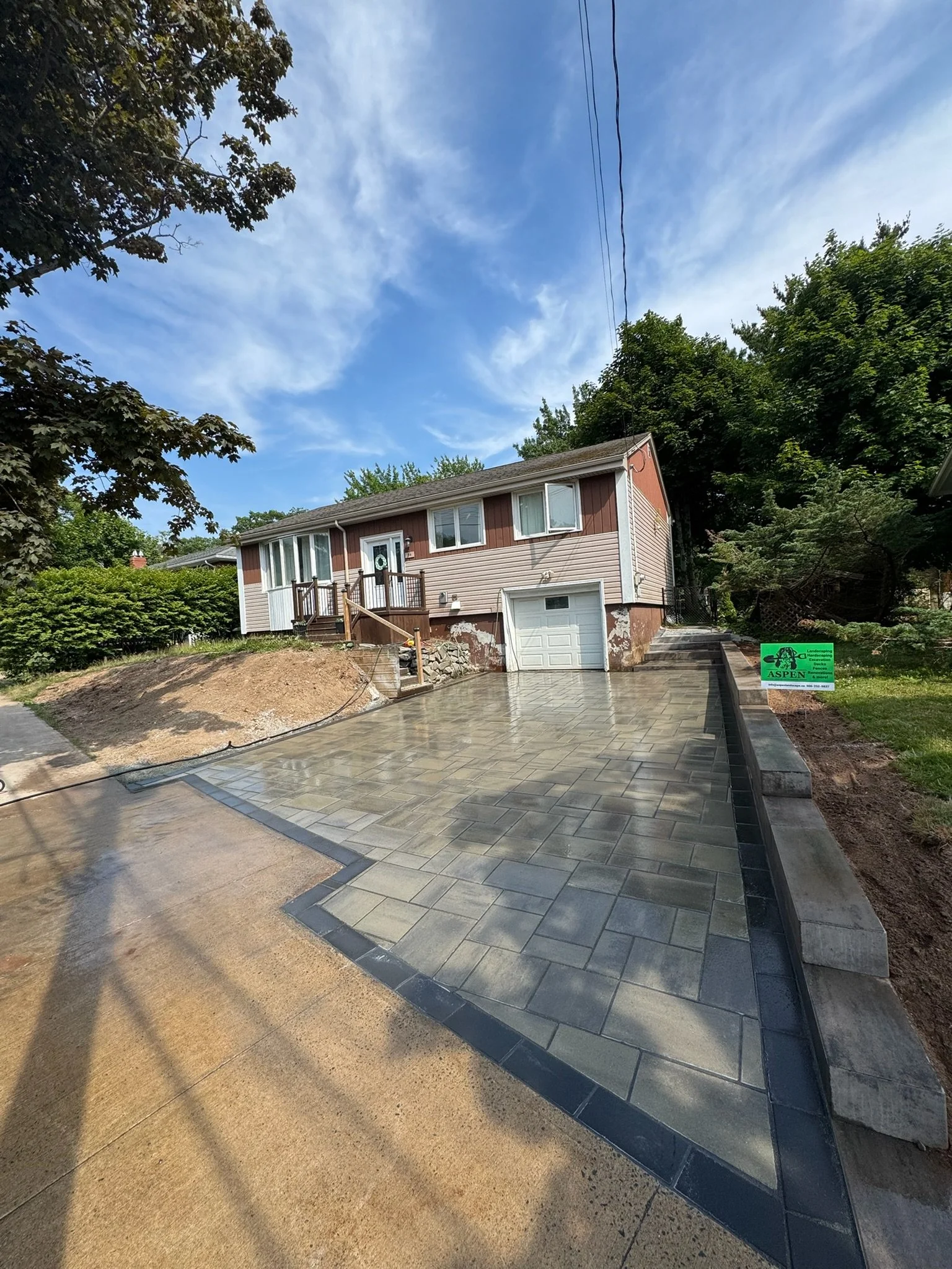 A house with a newly paved driveway, a garage door, and a front porch with steps leading up to the door. There is a green sign on the lawn that says 'Aspen' nearby, and trees surround the property under a blue sky with some clouds.