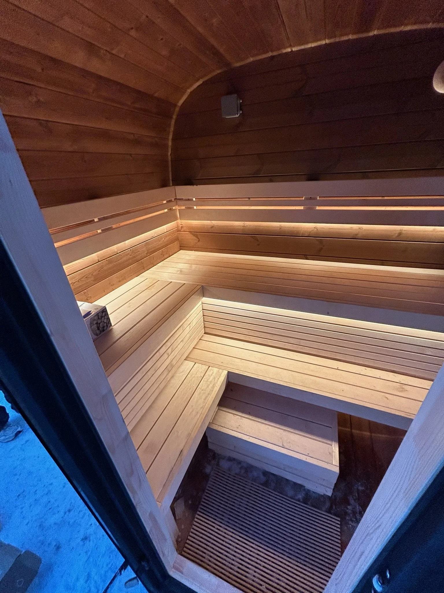 Interior of a small wooden sauna with tiered bench seating, a wooden floor, and wood-paneled walls and ceiling.