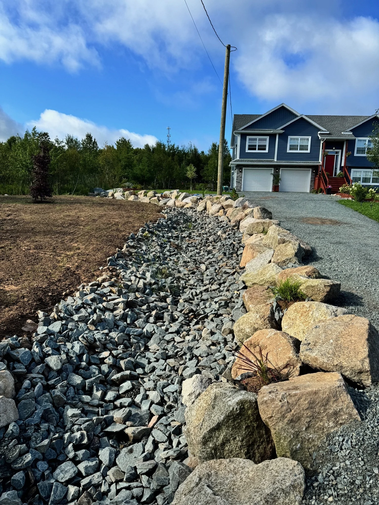 A gravel driveway with a large stone border leading to a blue house with a red staircase, in a rural setting with trees and a partly cloudy sky.