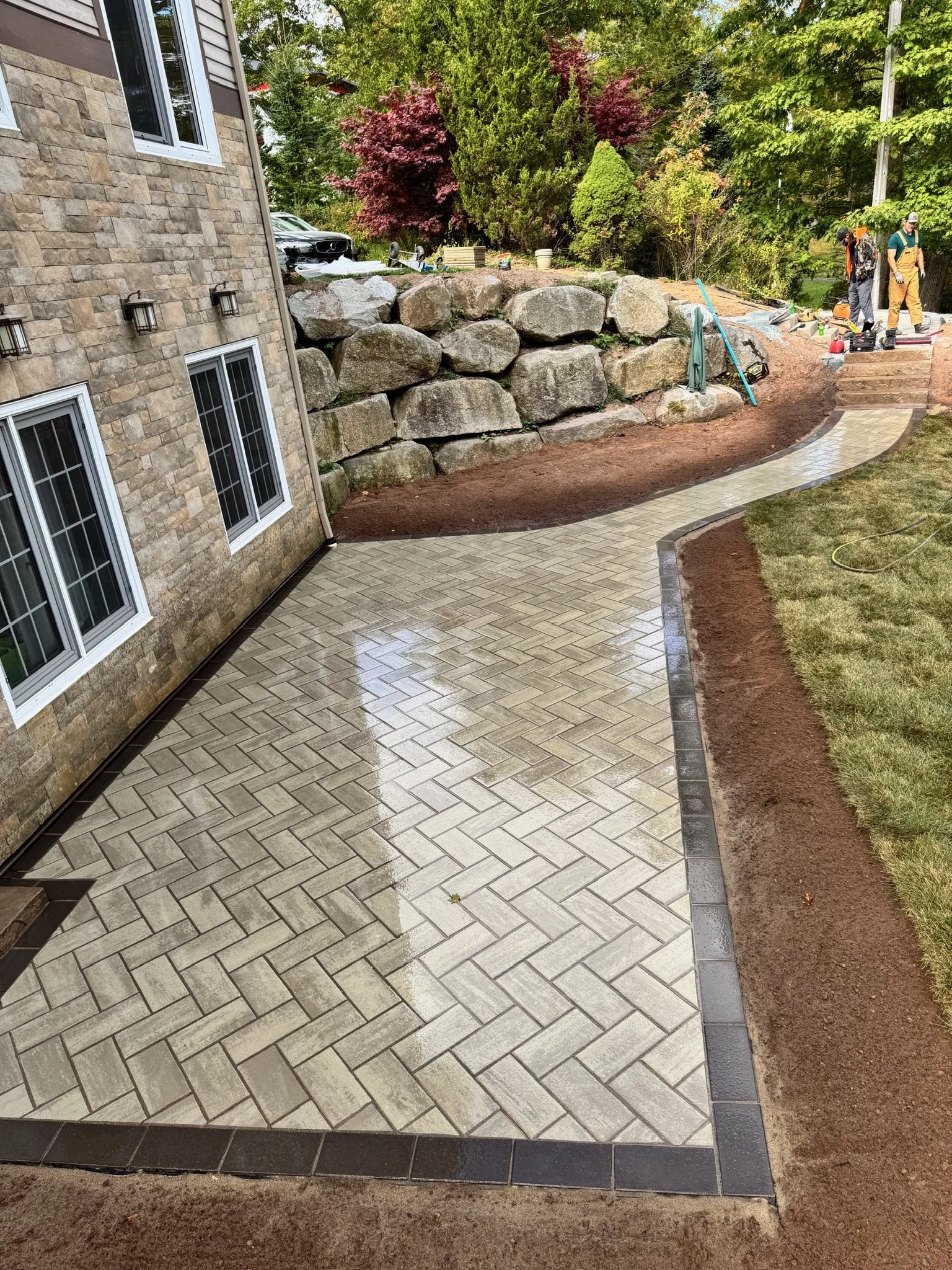 A newly paved, wet patio with interlocking brick pattern next to a stone house, with construction workers working in the background and a garden with large rocks and trees.