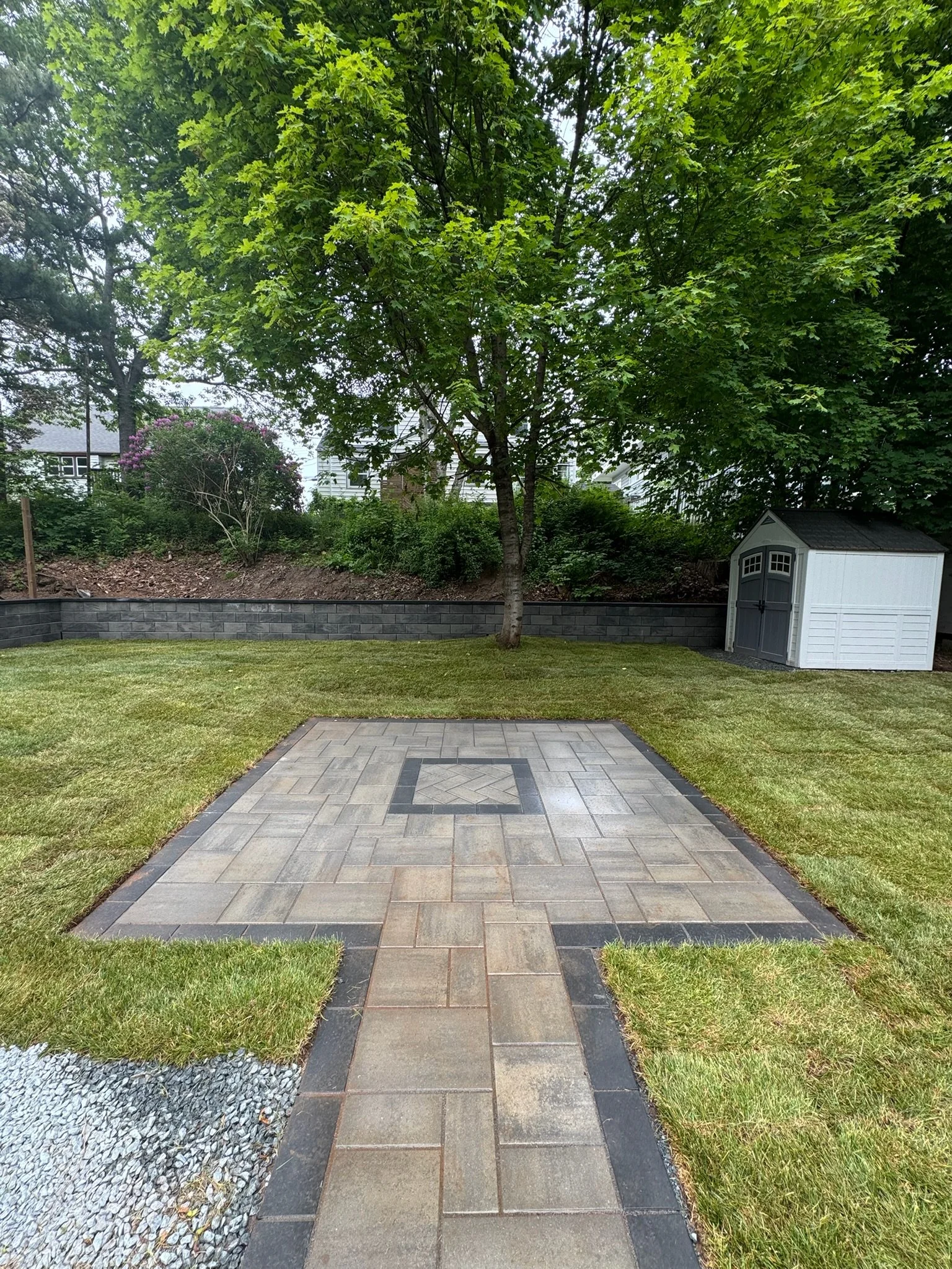 A backyard with a paved patio area with tan and dark gray bricks, surrounded by neatly trimmed grass, a large leafy tree, a white garden shed with gray doors, a lower retaining wall made of gray bricks, and a landscaped hillside with shrubs and flowe