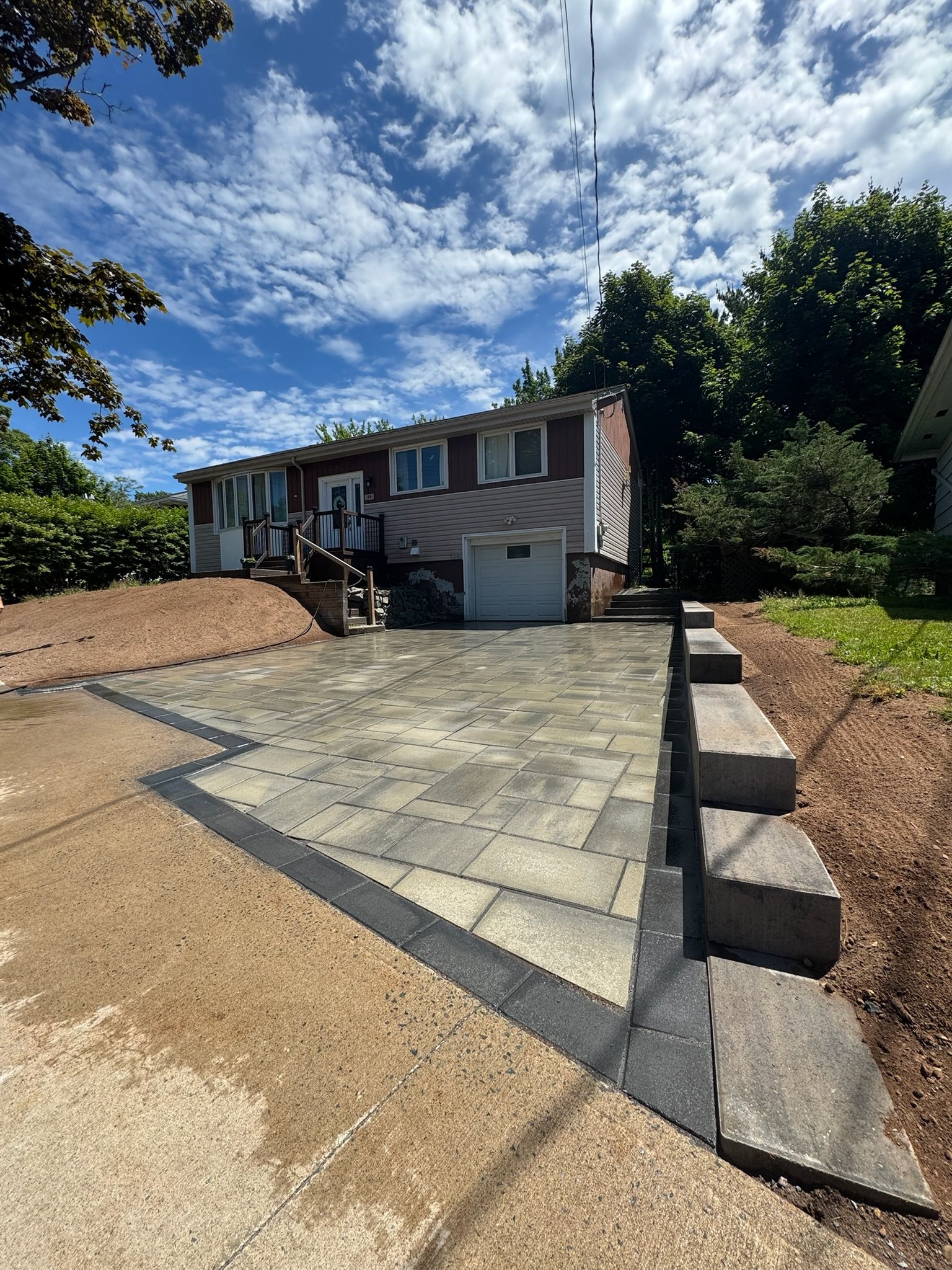 Newly paved driveway leading to a house with a garage, wooden steps, and a small porch, surrounded by trees and a partly cloudy sky