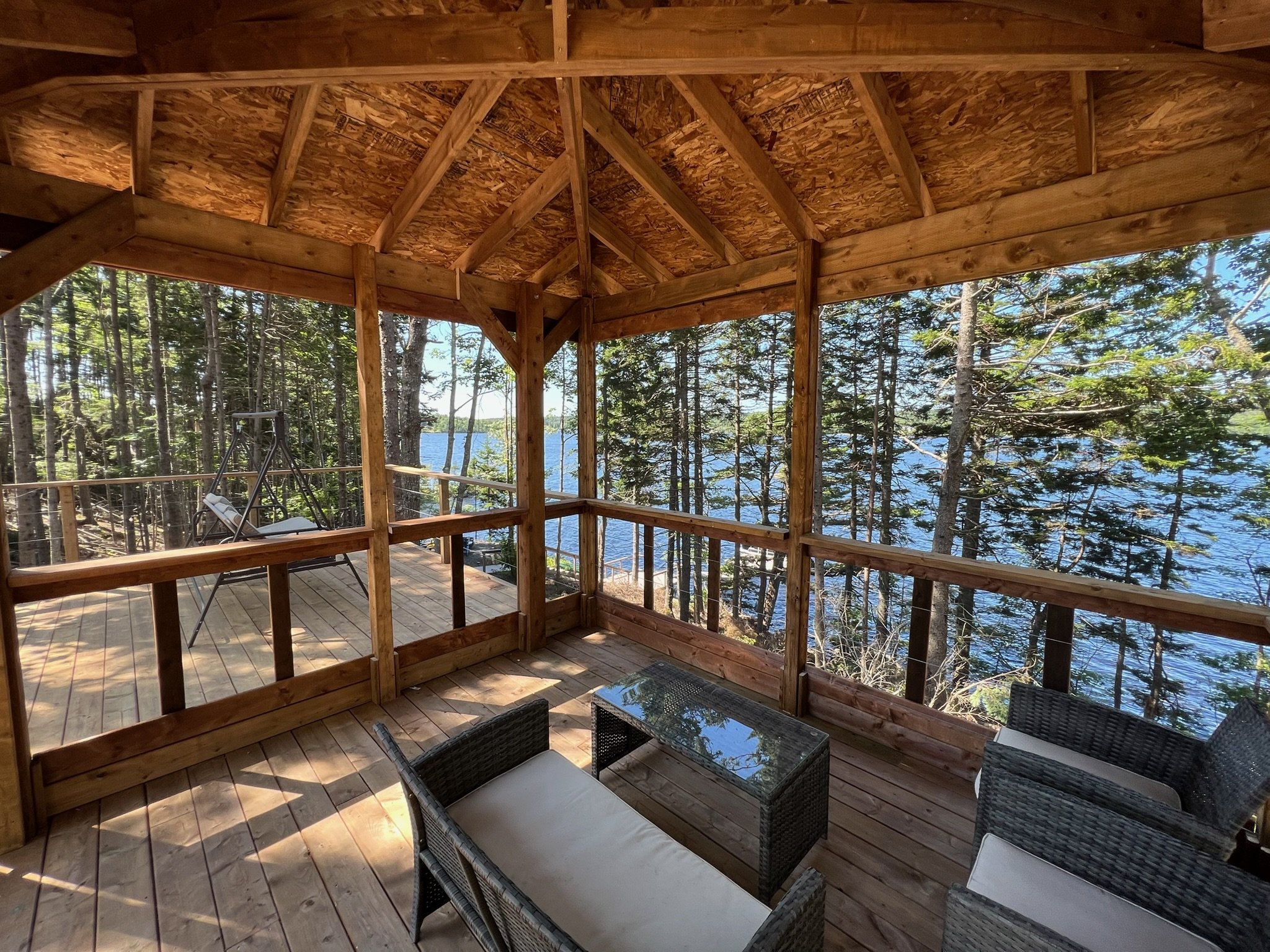 Wooden screened-in porch overlooking a lake with trees, furnished with wicker chairs and a glass-top table, with a swing on the deck outside.