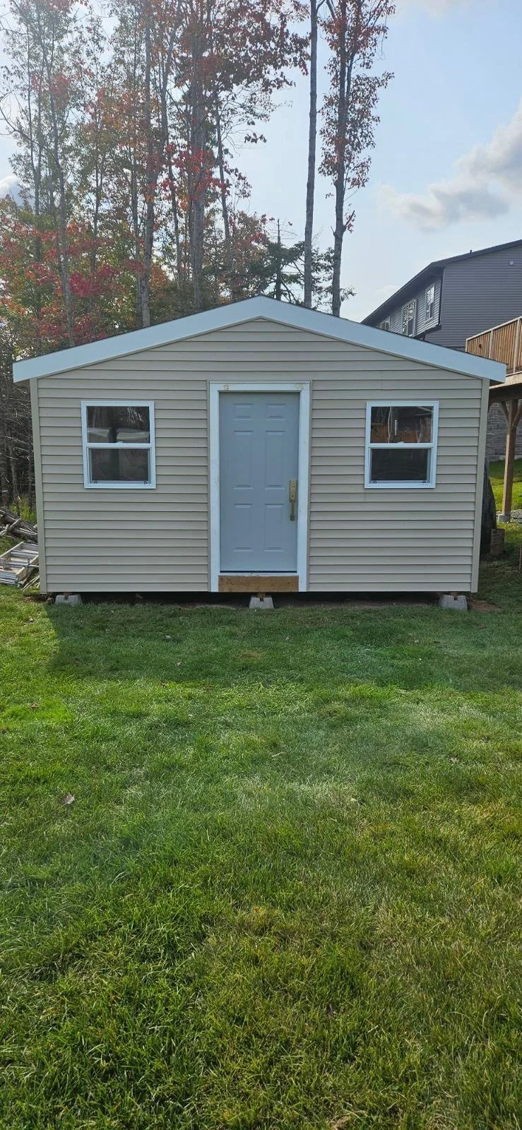 Small beige shed with a gray door and two windows, situated on a green lawn with trees and a neighboring house in the background.
