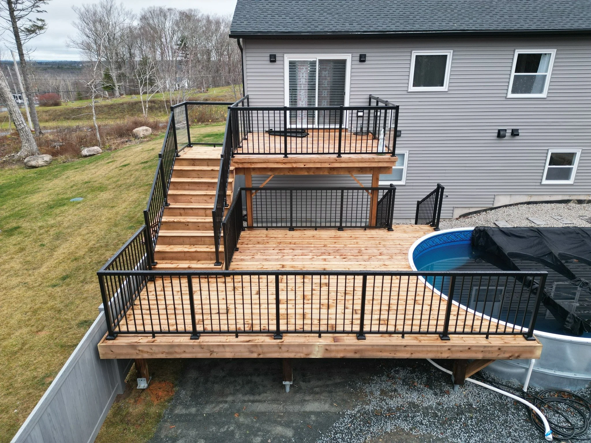 New multi-level wooden deck with black metal railings attached to back of gray house, with an above-ground pool partially covered nearby, and yard with grass and trees in background.