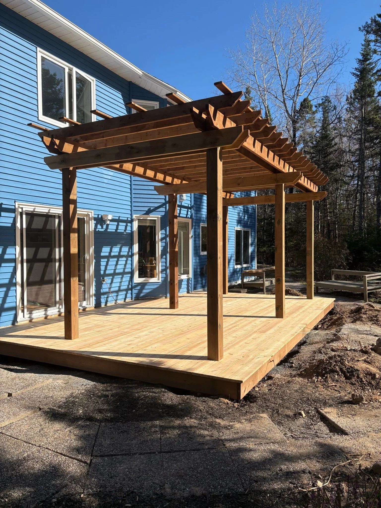 New wooden deck with a pergola being built outside a blue house, surrounded by trees and clear blue sky.