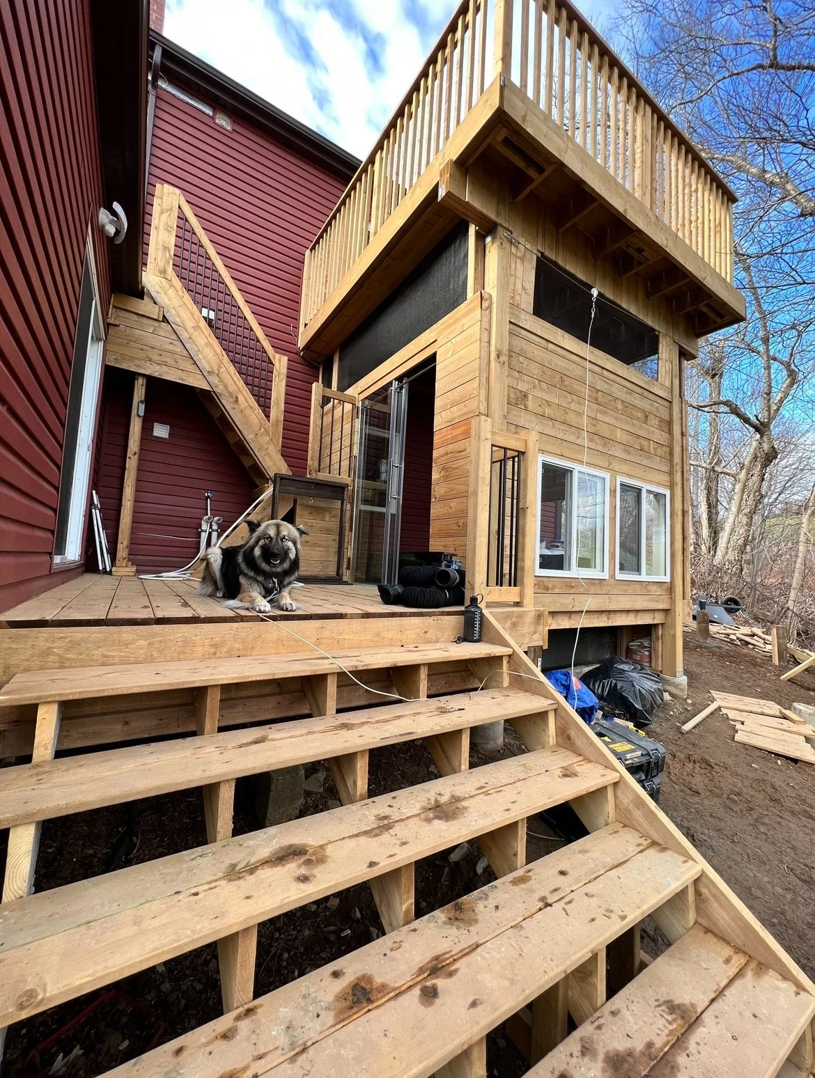 A partially constructed wooden deck with stairs leading up to a new two-story house, a dog sitting on the deck, and construction materials around the site.