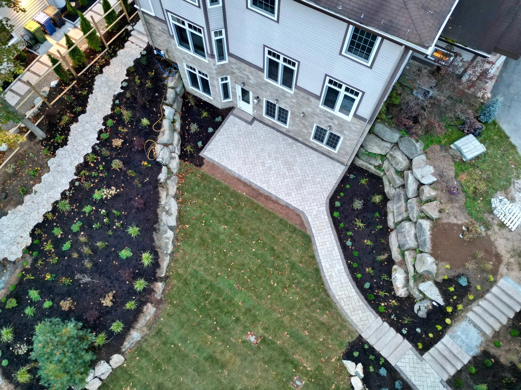 Aerial view of a backyard with a house, landscaped garden beds, a curved stone pathway, stone steps, and a patio area.
