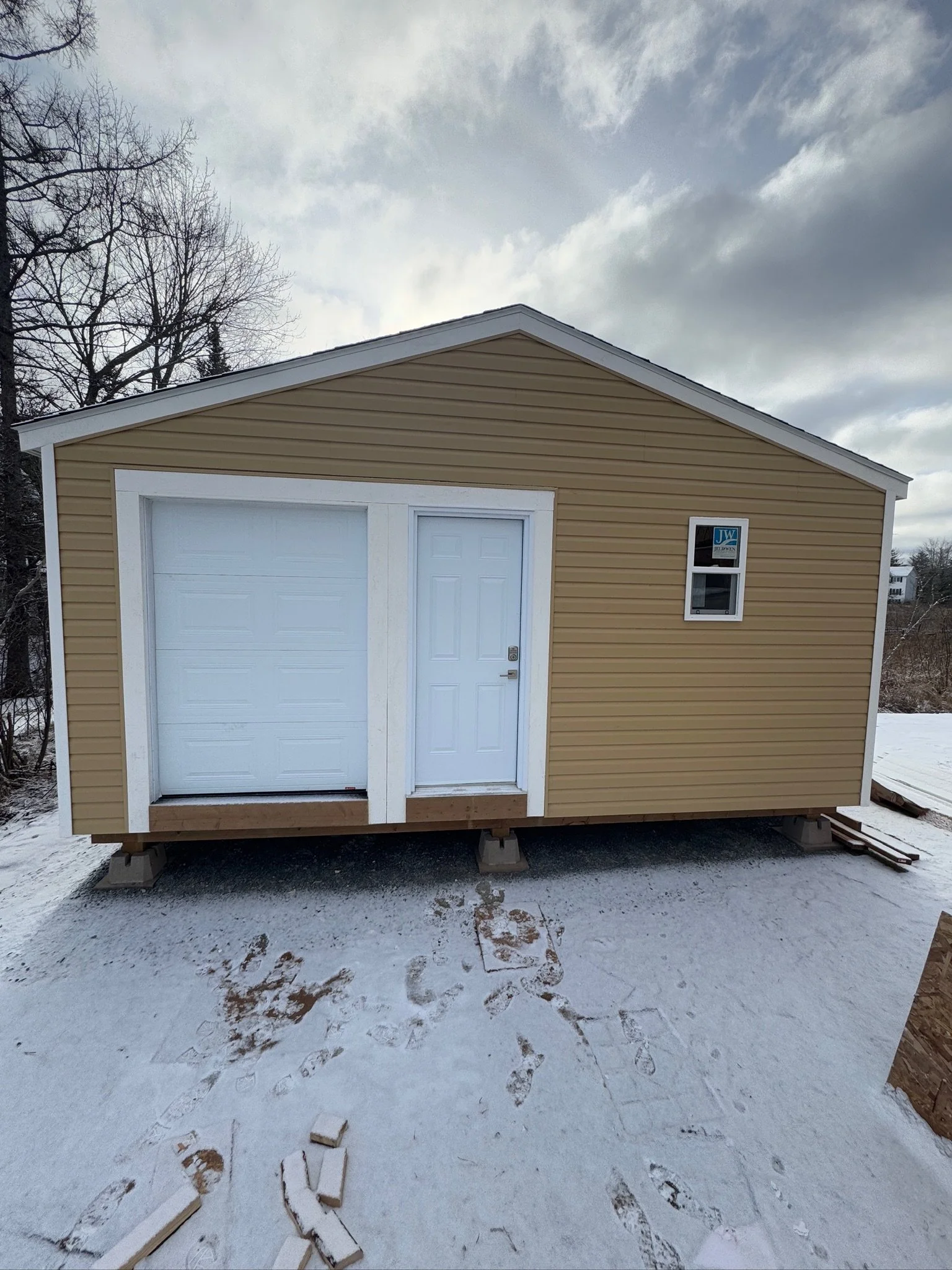 A small beige garage with white trim and a white door, elevated on concrete blocks, with snow on the ground and a cloudy sky overhead.